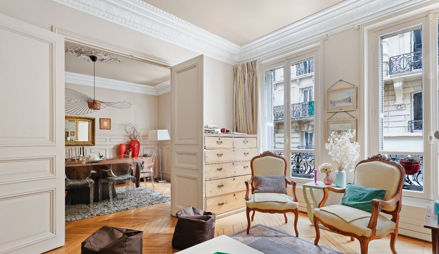 Salle à manger haussmannienne d'un appartement meublé rue Lentonnet Paris 9e, suspension Vertigo au-dessus d'une table ovale et chaises Louis Ghost sous des moulures d'époque