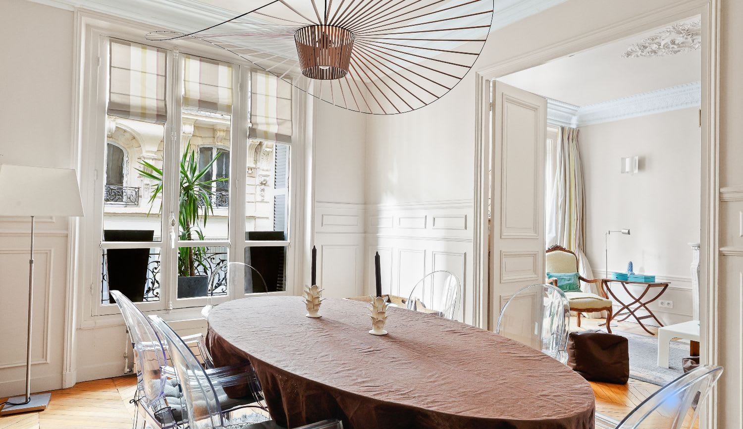 Salle à manger haussmannienne d'un appartement meublé rue Lentonnet Paris 9e, suspension Vertigo au-dessus d'une table ovale et chaises Louis Ghost sous des moulures d'époque