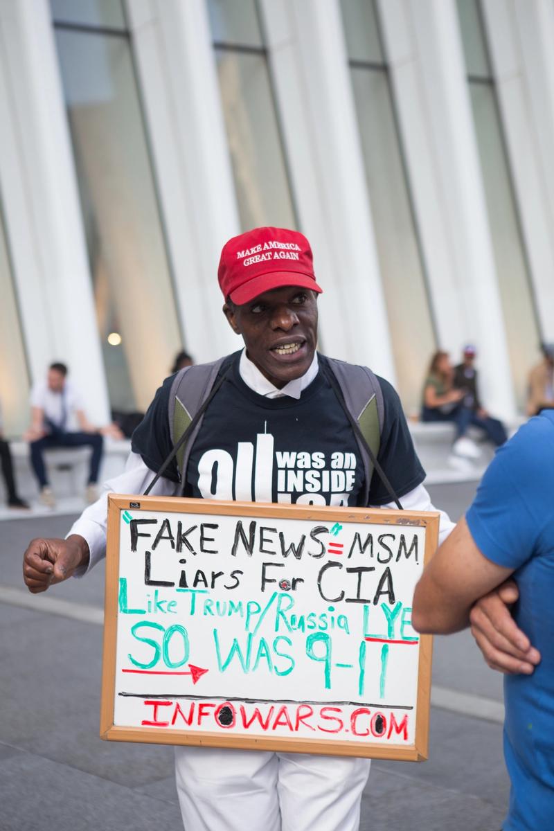 En mann med en caps som har teksten "Make America great again" står på gaten med et skilt rundt halsen. På skiltet står det ""FAKE NEWS" = MSM. Liars for CIA. Like Trump/Russia LYE So was 9-11. INFOWARS.COM" 
