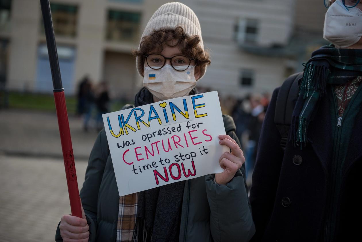 En person med lue og munnbind holder et skilt der det står "Ukraine was opressed for CENTURIES. Time to stop it NOW" Ukraine er skrevet med et hejrte over I-en og med blått og gult.