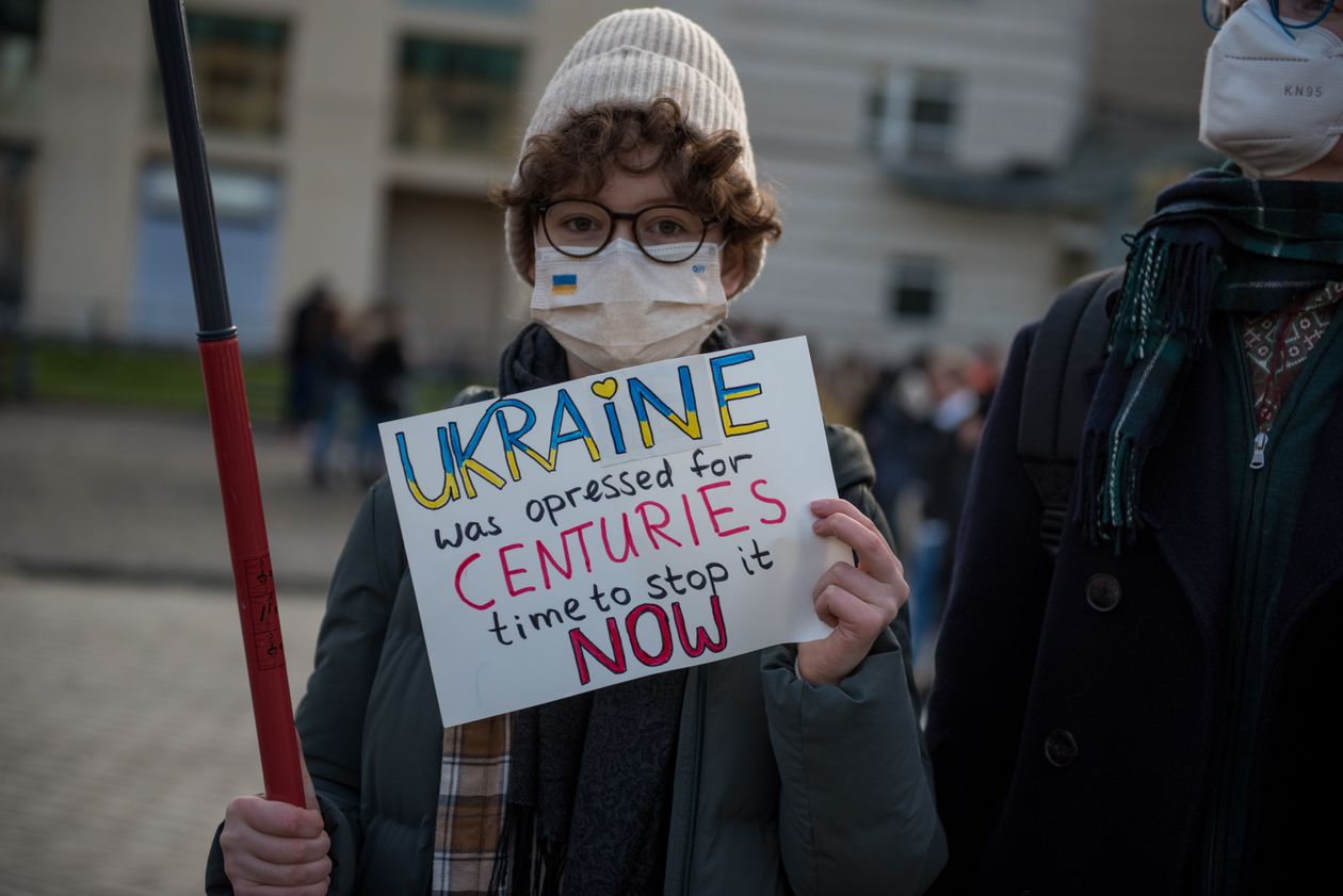 En person med lue og munnbind holder et skilt der det står "Ukraine was opressed for CENTURIES. Time to stop it NOW" Ukraine er skrevet med et hejrte over I-en og med blått og gult.