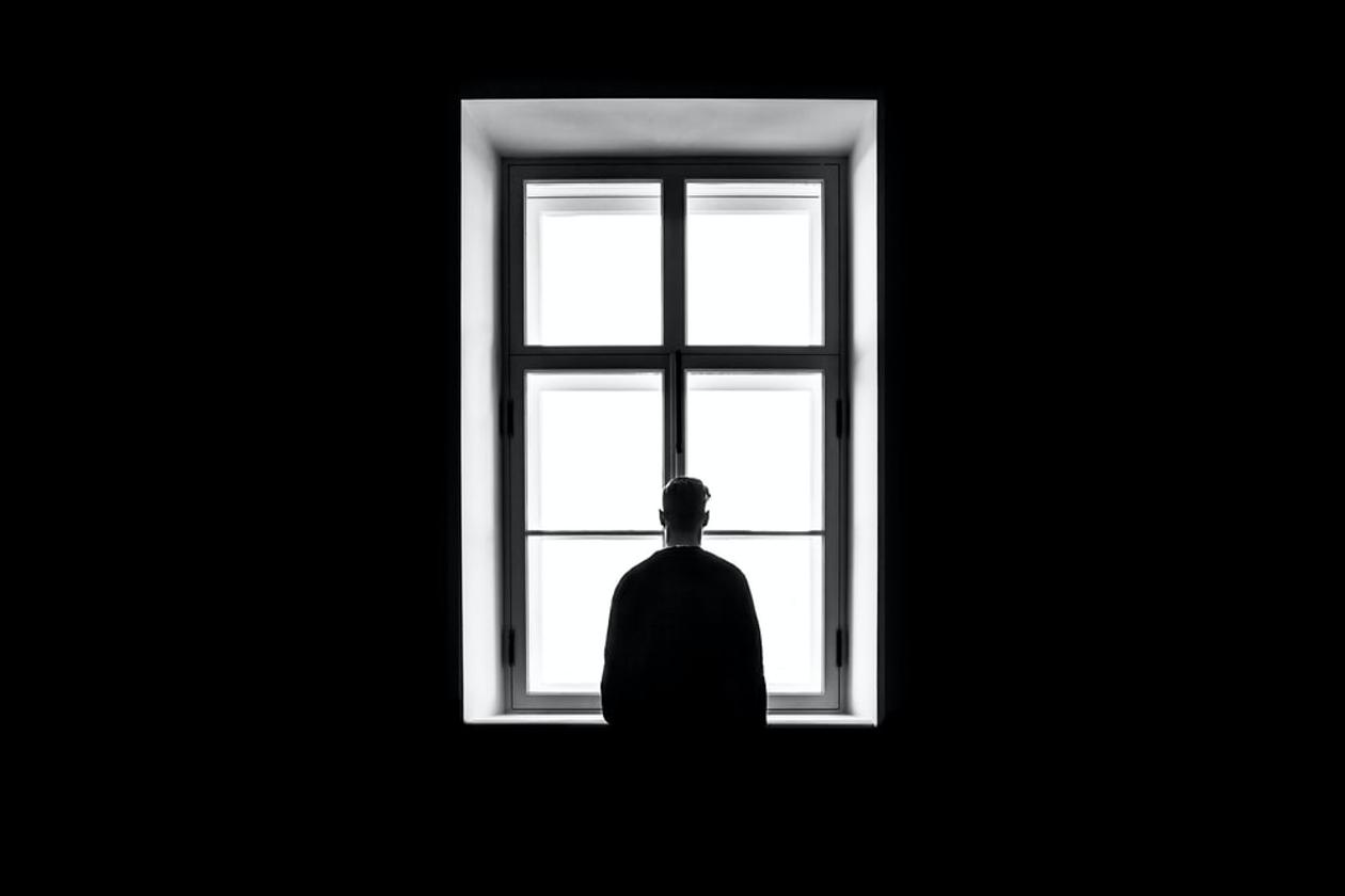 Et svart rom med et stort vindu. En mann står med ryggen til og ser ut av vinduet.