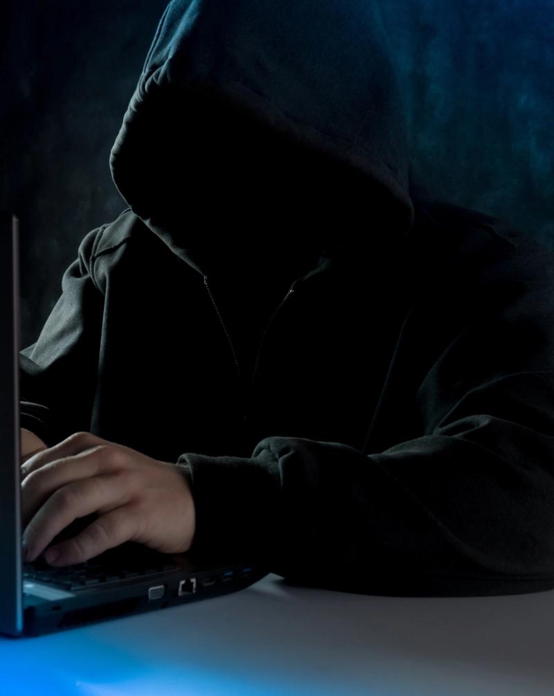 Et mørkt bilde av en person foran en laptop. Personen har et hette som skjuler ansiktet.