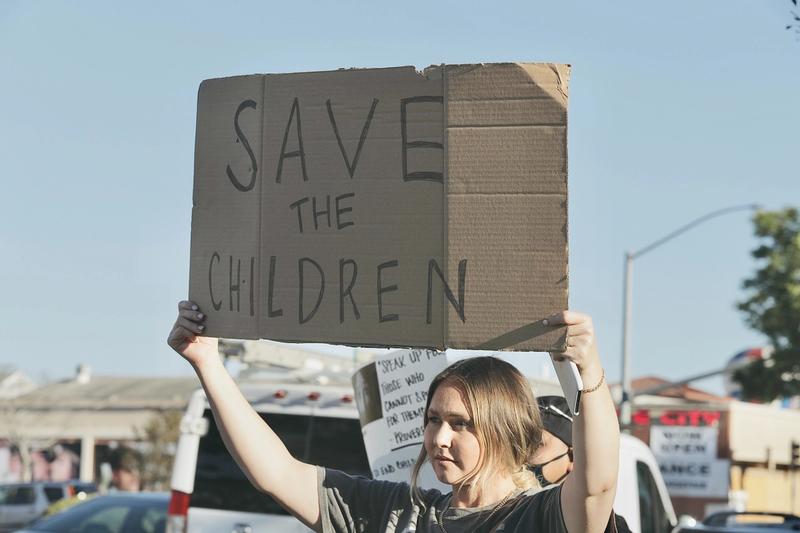 En kvinne er på en demonstrasjon og holder opp en papp-plakat der hun har skrevet "SAVE THE CHILDREN". Bak budskapet ligger en konspirasjonsteori om at en pedofil elite styrer USA.