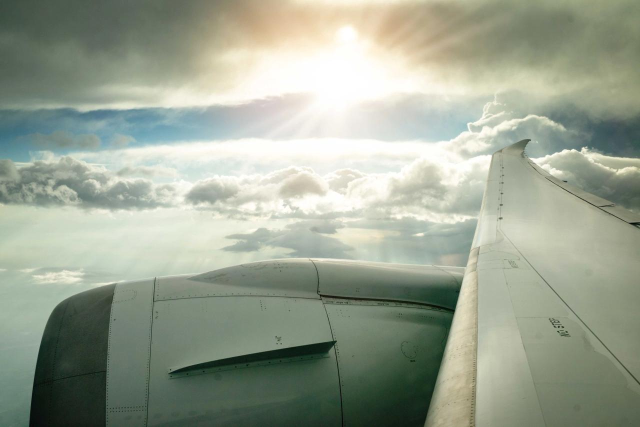 An airplane wing seen from a passenger window, above a layer of clouds with the sun shining through.
