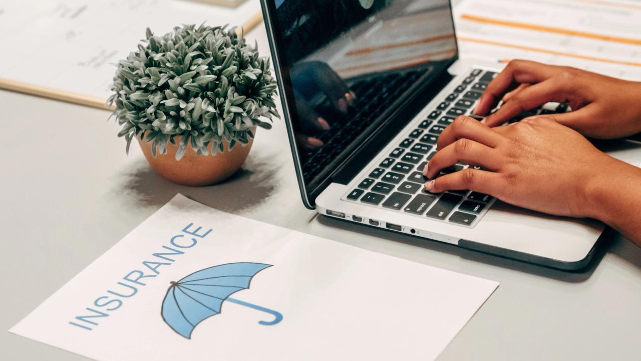 A person typing on a laptop at a white desk, beside a printed sheet headed Insurance with a blue umbrella illustration.