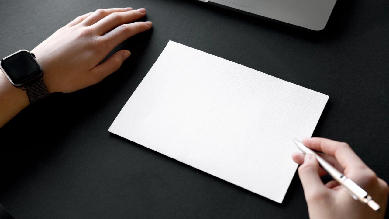 A hand holding a pen over blank paper beside a laptop.