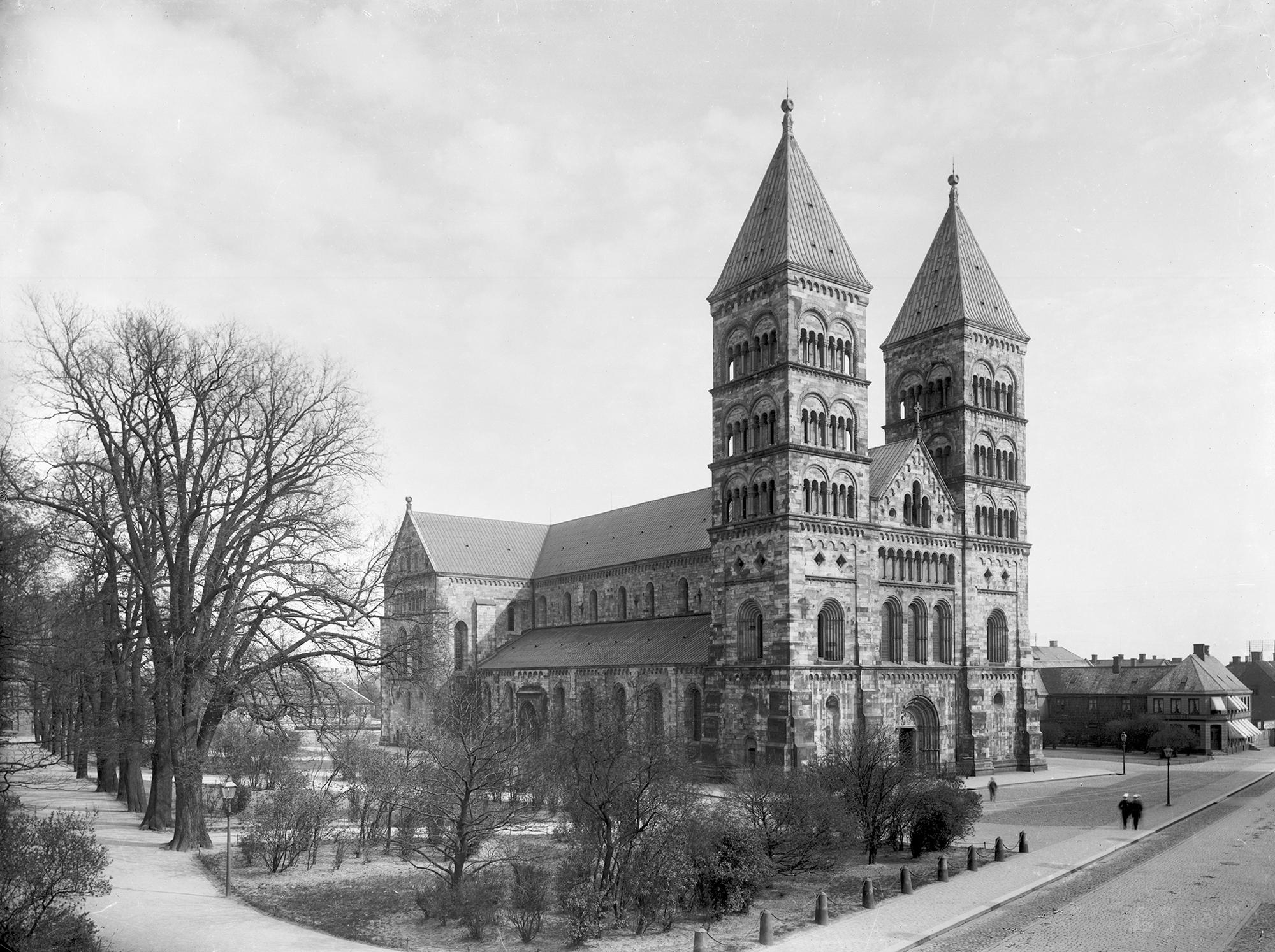 Black and white photo of a grand stone cathedral with two prominent square towers, next to a park with bare trees and a street.