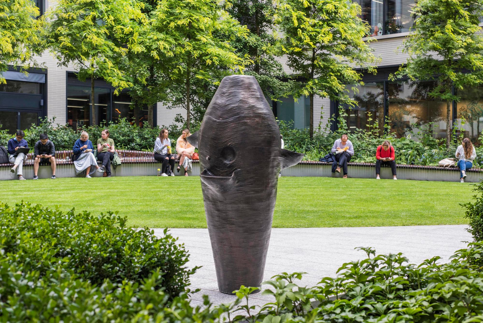 A tall, bronze-colored abstract sculpture stands in a green park, with people relaxing on benches and trees in the background.