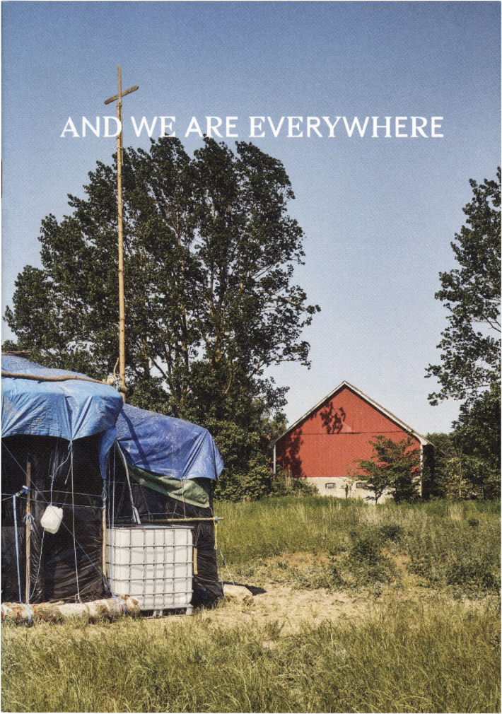 Makeshift structures covered in blue tarps, with a tall cross on a pole in a grassy field. A red barn stands in the background. Text reads: 'AND WE ARE EVERYWHERE'.