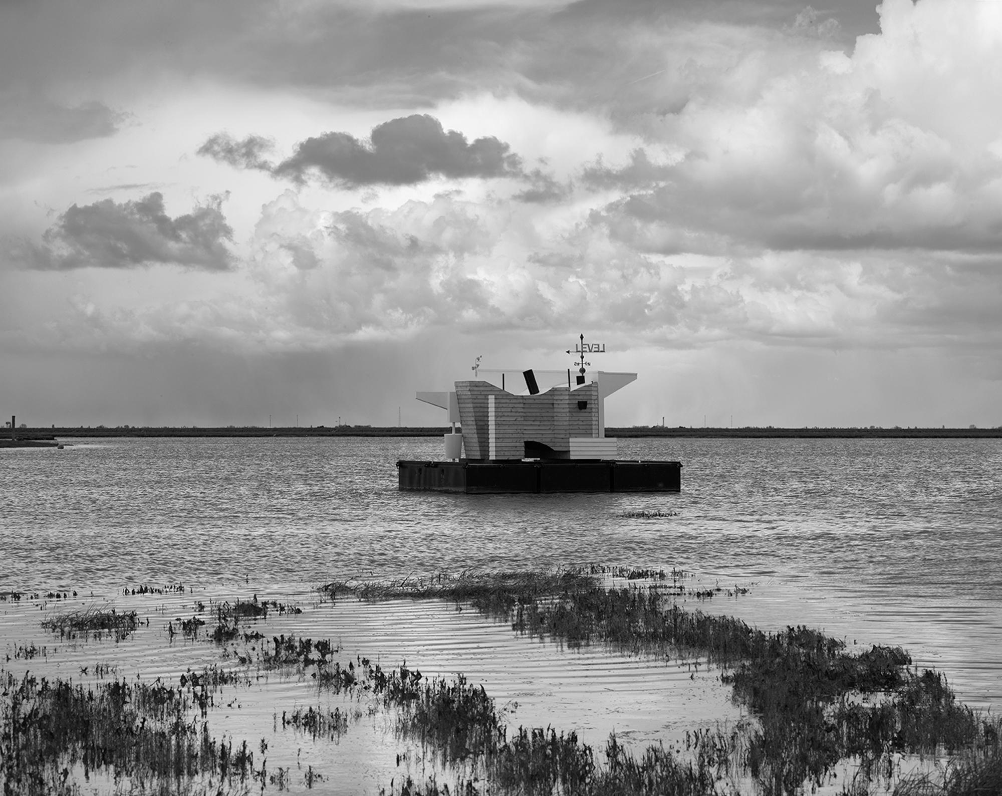 Black and white image of a geometric structure floating on water beneath a cloudy sky.