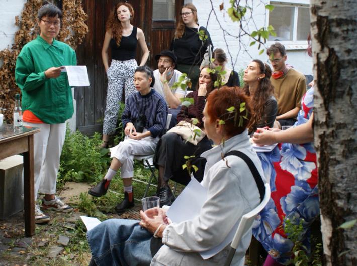 A diverse group of people gathered outdoors, listening to a speaker holding papers.