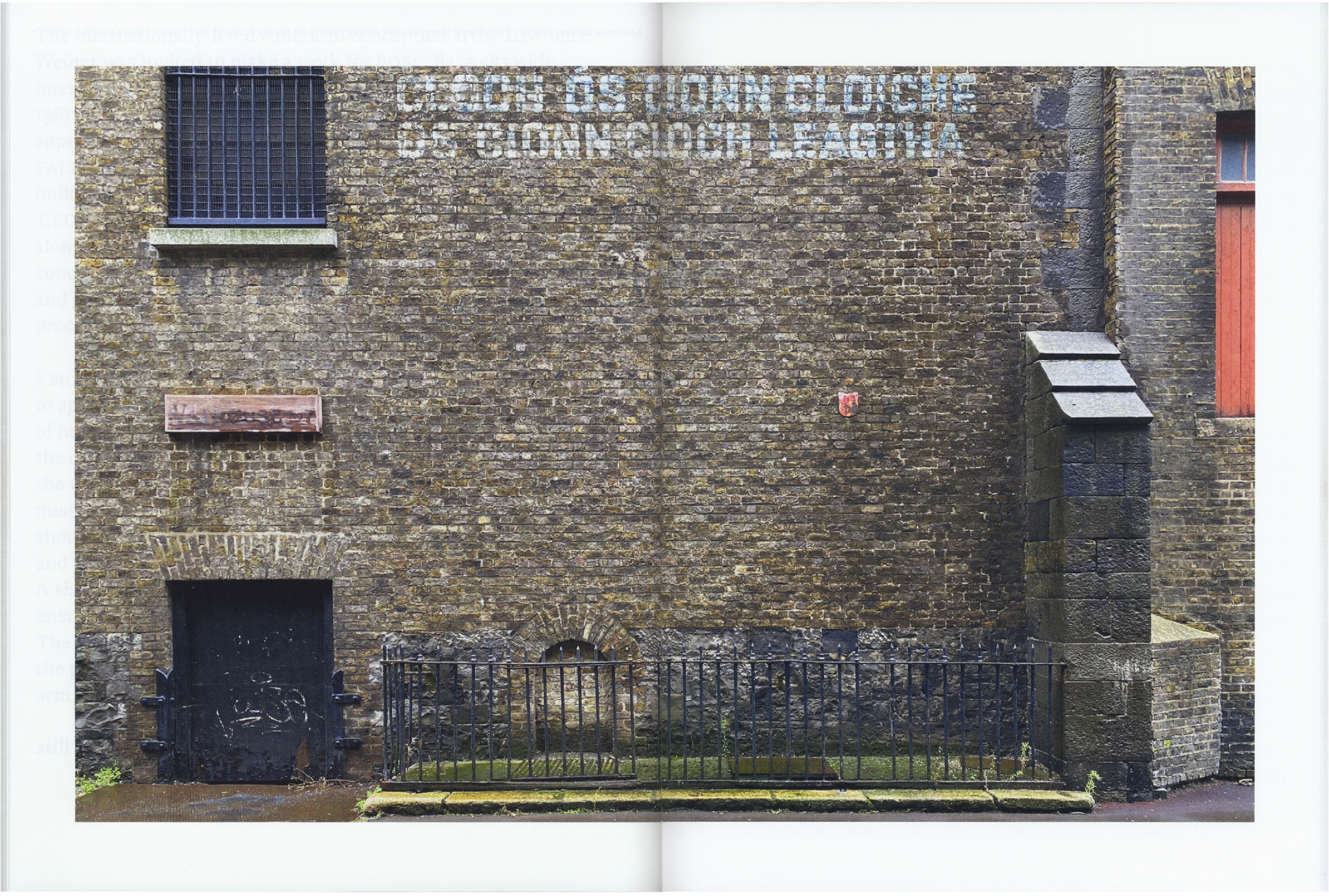 A weathered brick wall with a barred window, an arched doorway, faded painted text, and an iron fence.