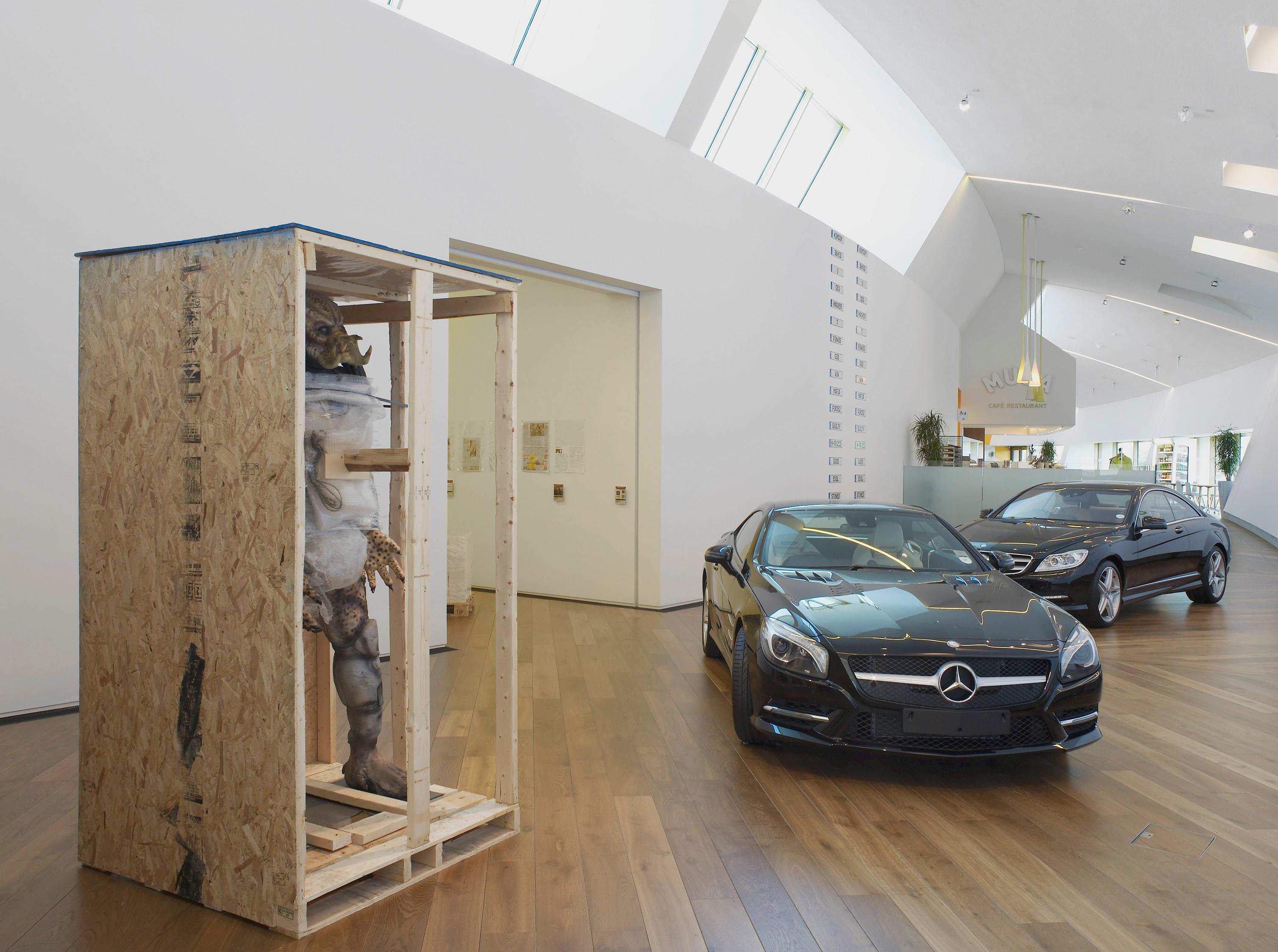 A modern showroom features a tall wooden crate with a wrapped humanoid figure on the left, and two black Mercedes-Benz cars on the right.