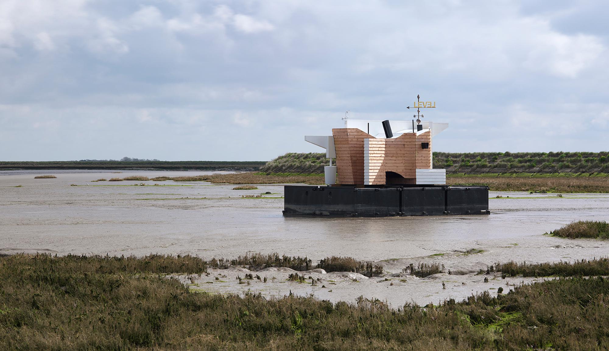 A modern wood and white floating structure sits in a muddy estuary surrounded by marshland under a cloudy sky.