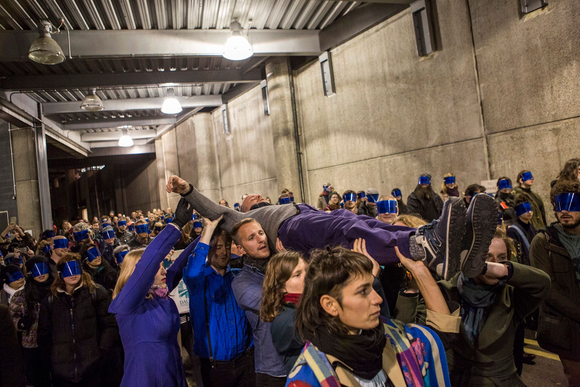 A crowd of people, many wearing blue blindfolds, carry a man aloft through a concrete corridor.