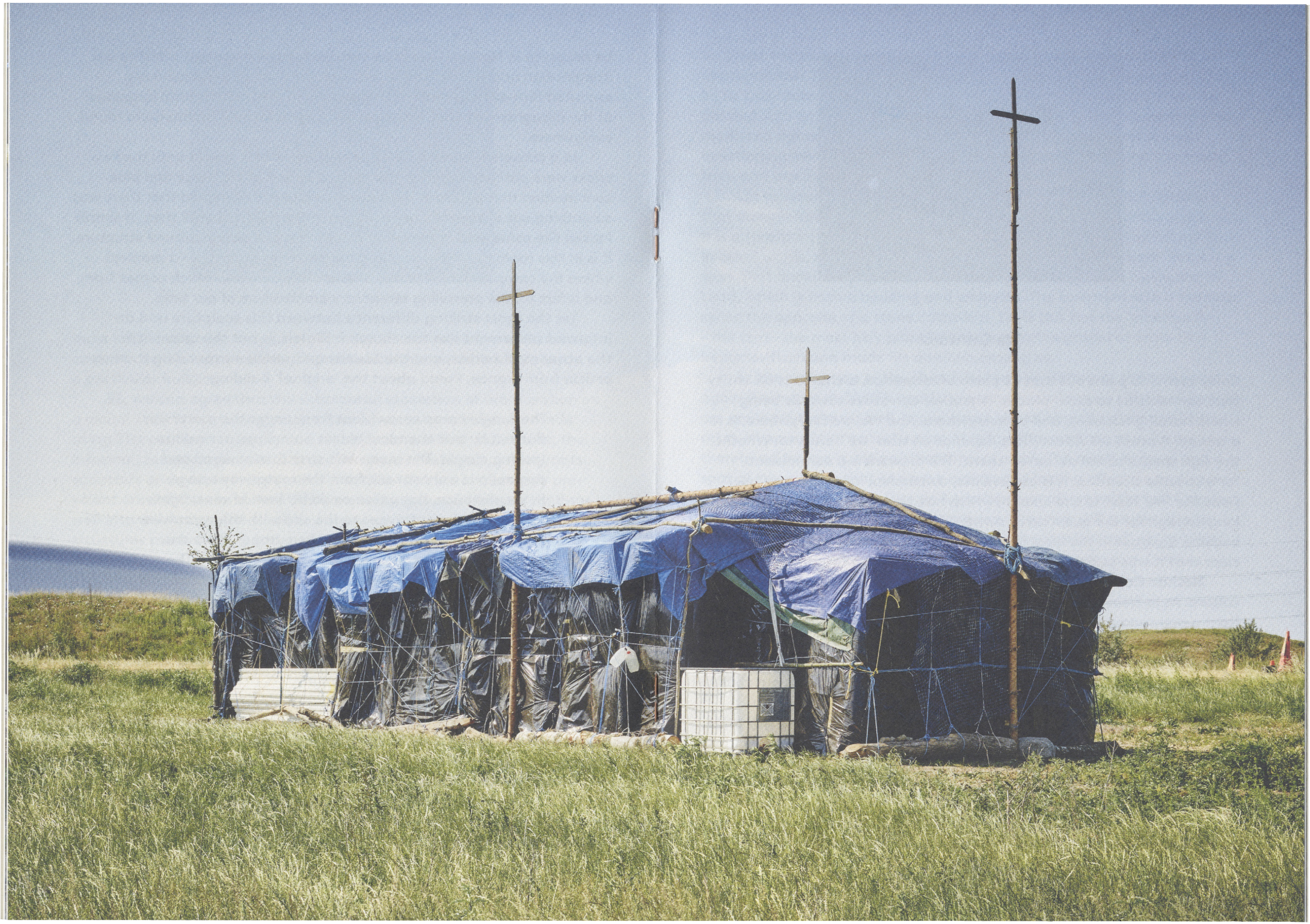 A makeshift church constructed from blue and black tarps with three crosses on top, situated in a green field.