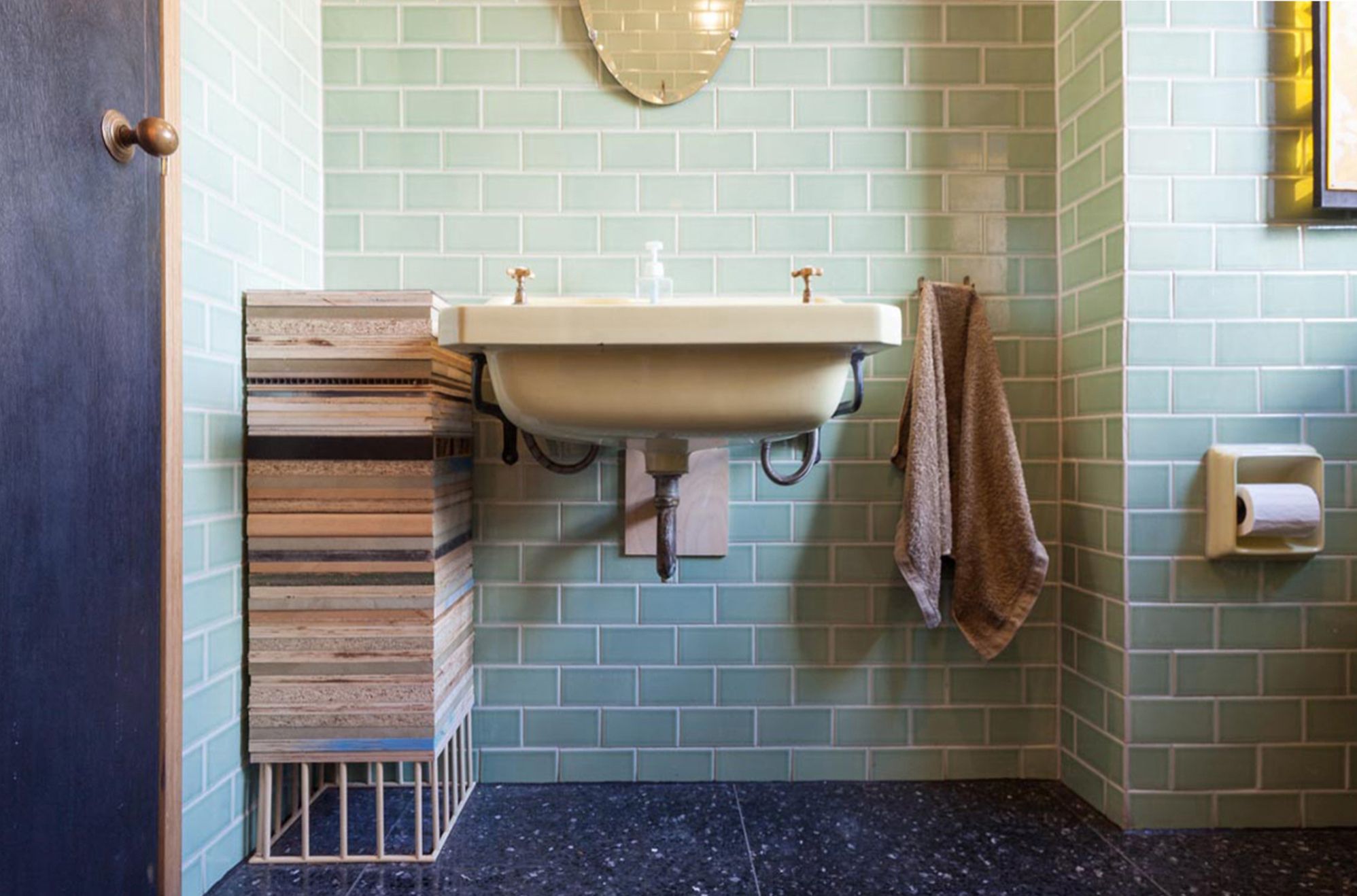 Bathroom with light green subway tiles, a cream wall-mounted sink, a tall layered plywood cabinet, and a dark speckled floor.