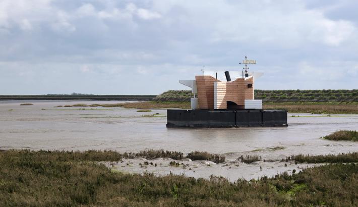 A modern, partially wooden building on a floating platform in a muddy marsh.
