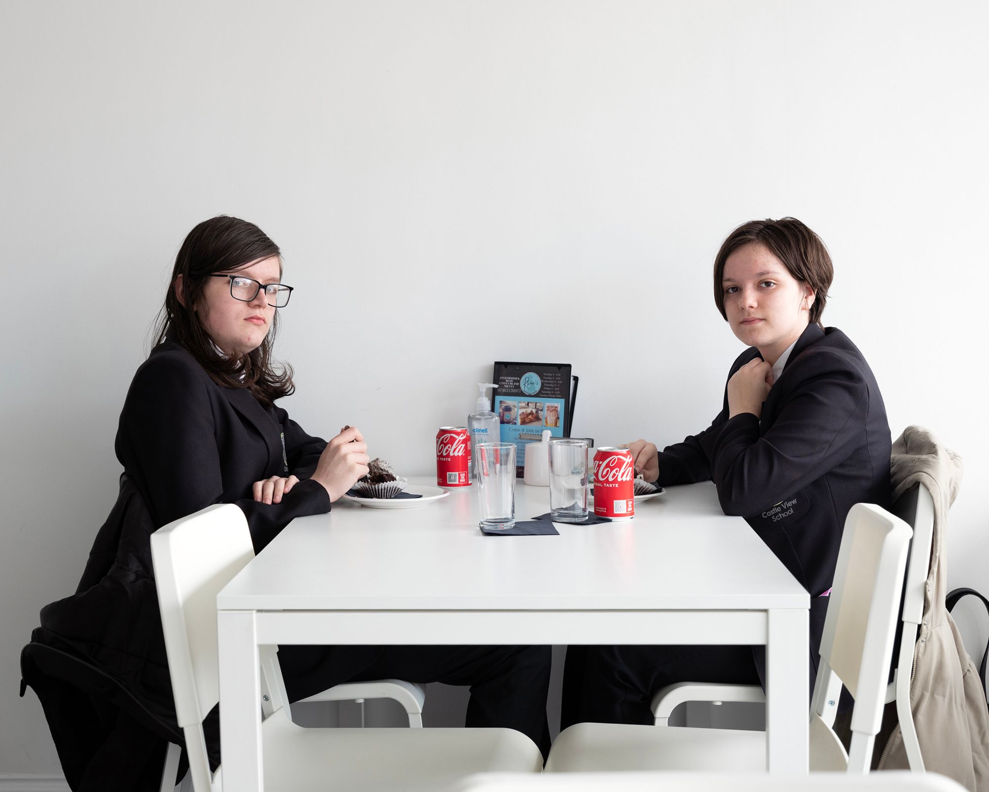 Two teenagers in dark blazers sit at a white table with cake and Coca-Cola cans.