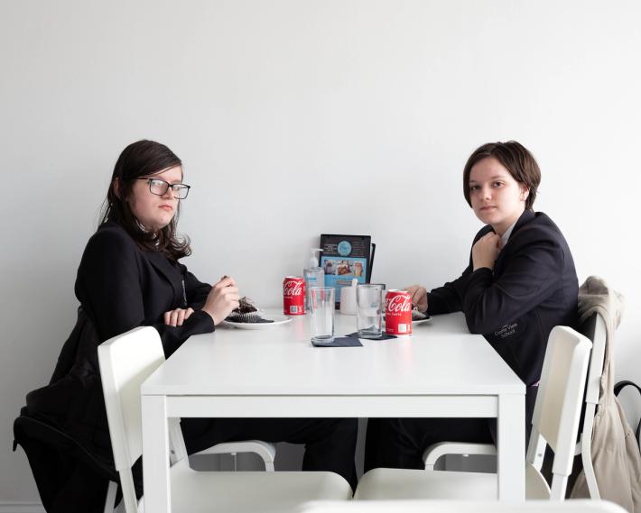 Two teenagers in dark blazers sit at a white table with cake and Coca-Cola cans.