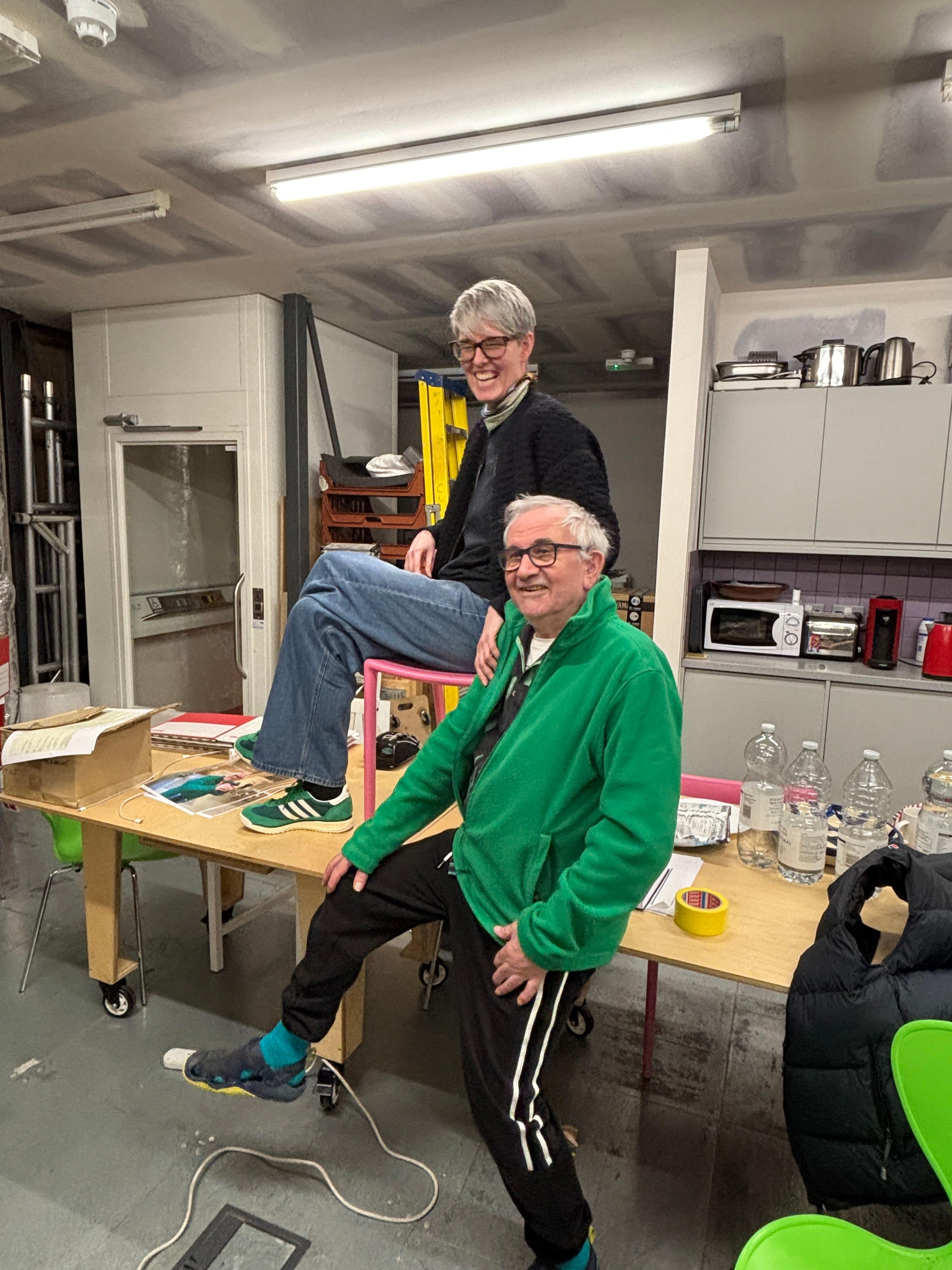 Two smiling people in a playful pose; a woman sits on a table while a man leans against it with one leg raised.