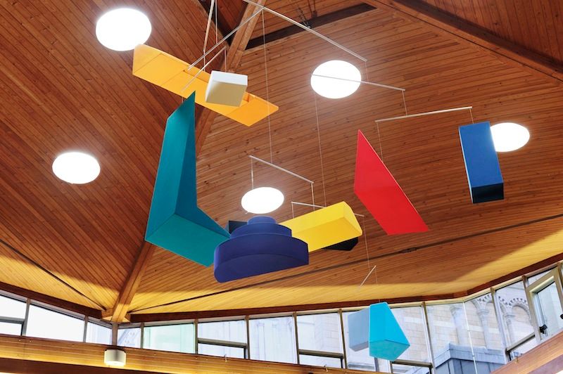 Abstract mobile with colorful geometric shapes hangs from a wooden vaulted ceiling with circular lights.