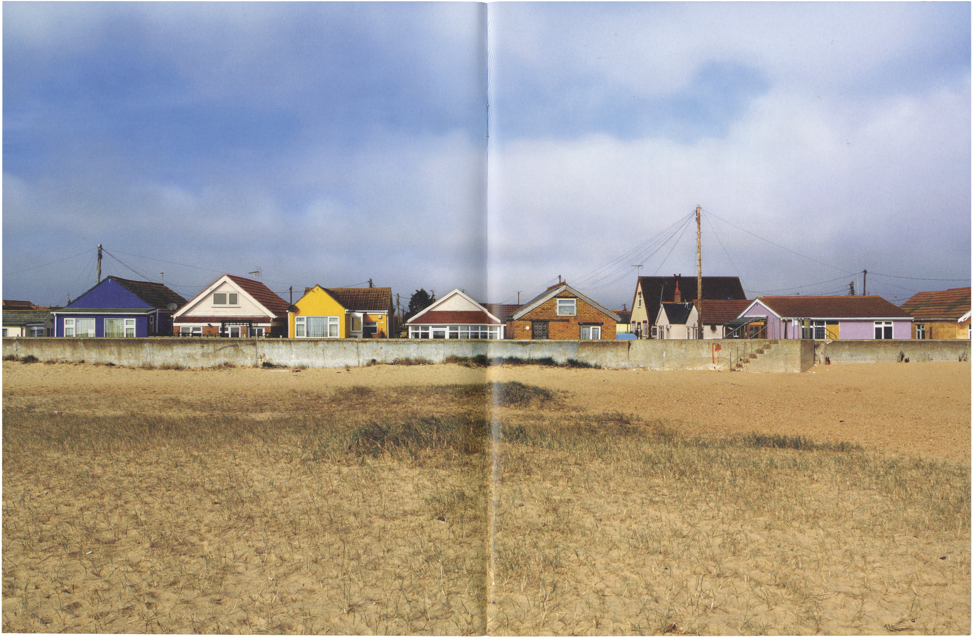 A row of colorful houses behind a concrete sea wall, viewed from a sandy beach under a partly cloudy sky.