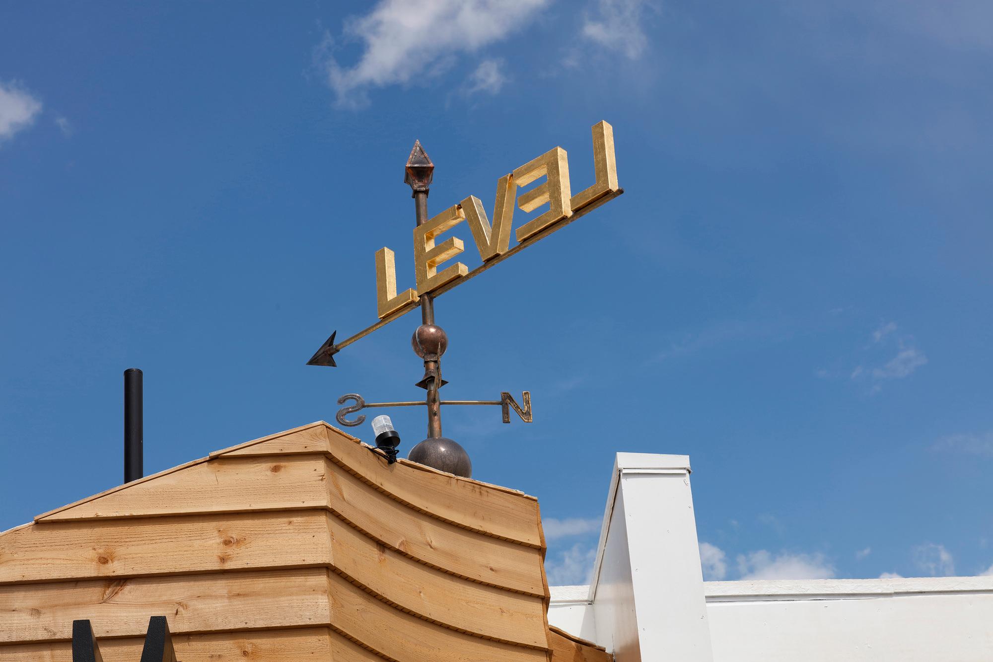 A weathervane with gold "LEVEL" letters on a wooden rooftop against a blue sky.