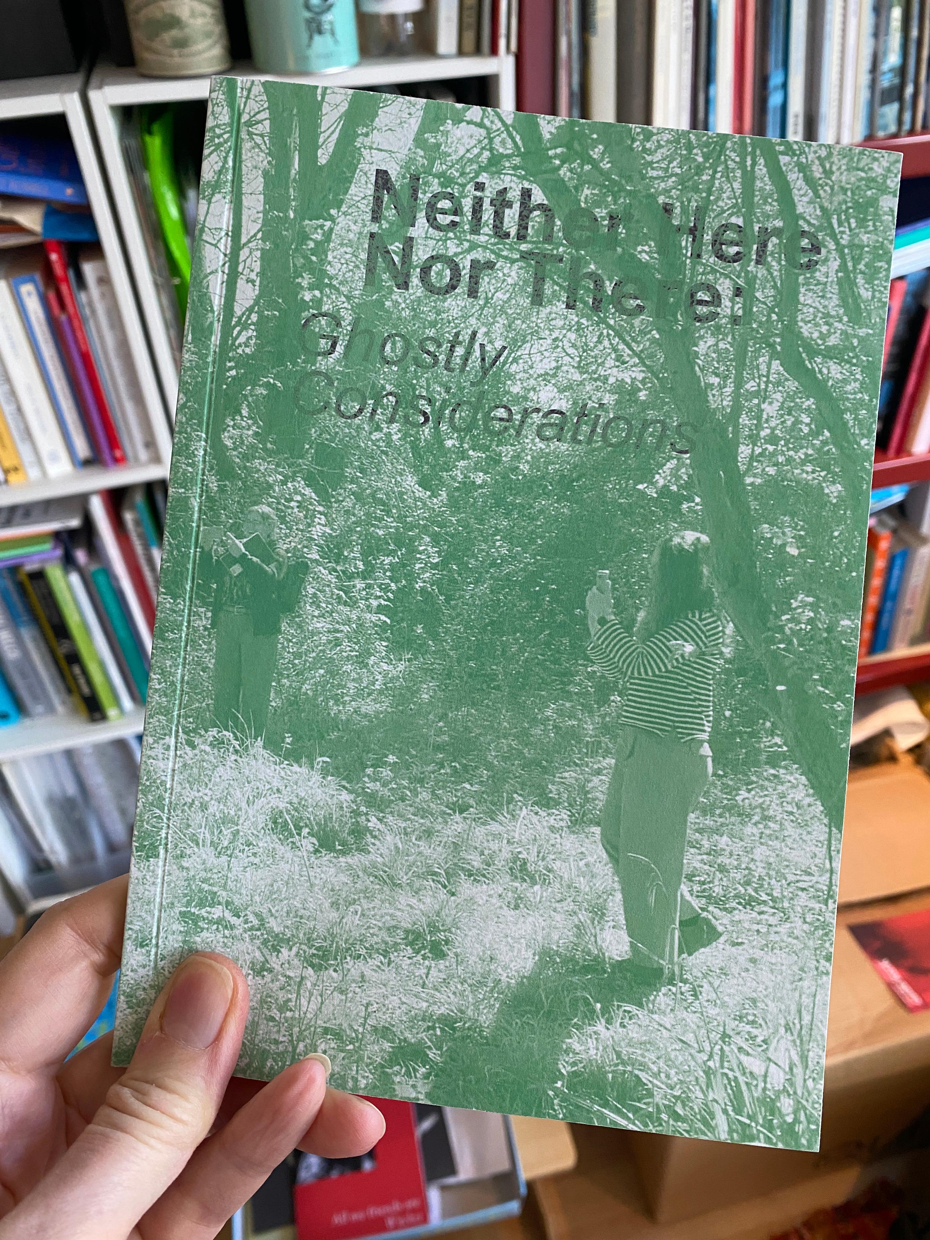 A hand holds a green book titled "Neither Here Nor There: Ghostly Considerations," featuring a faint, forest-like image with two figures.