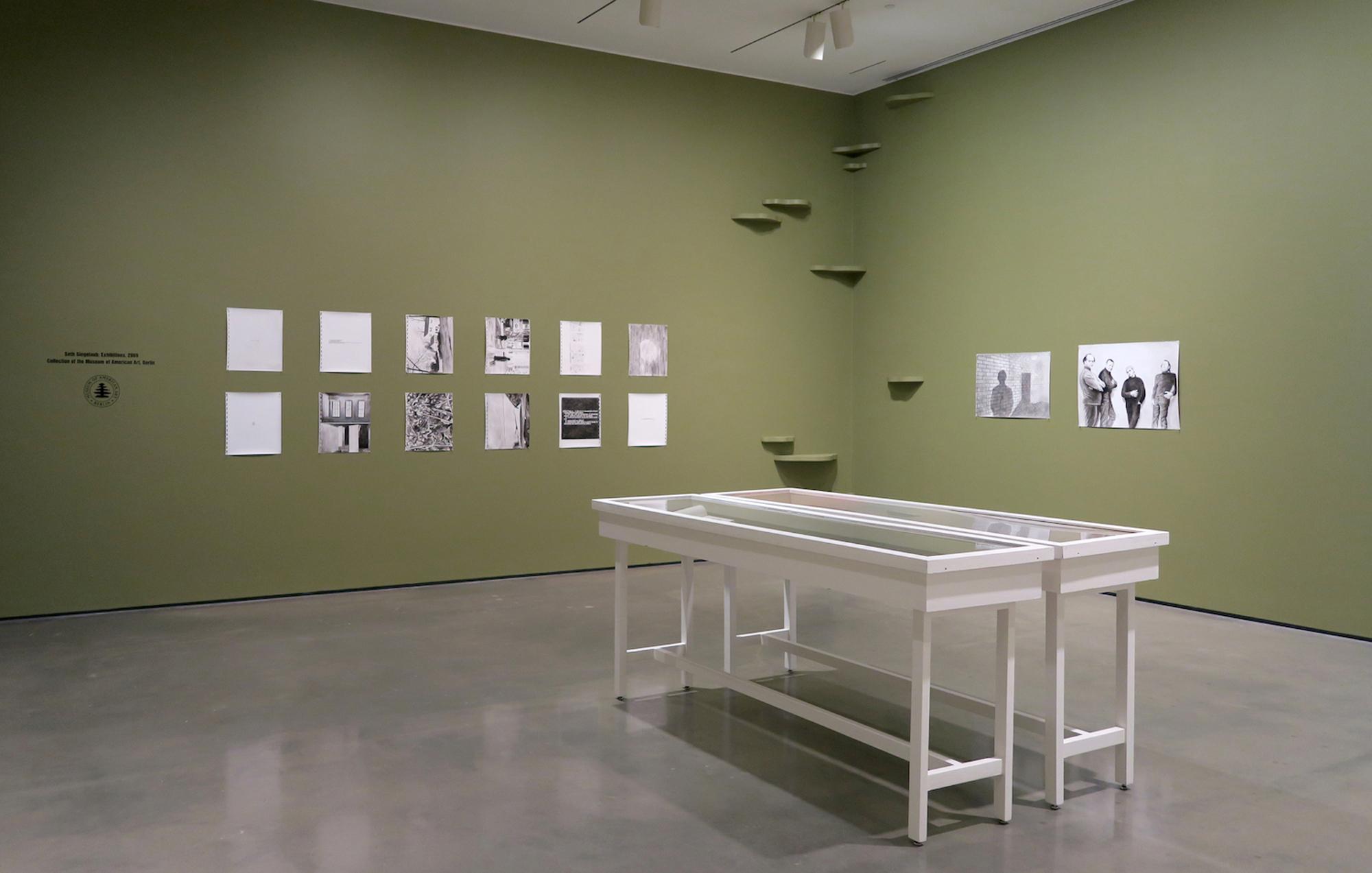 Art gallery with olive green walls, concrete floor, displaying a grid of 12 small black and white works and a text inscription on the left, two large black and white photos and ascending green shelves on the right, and two white display cases in the foreground.