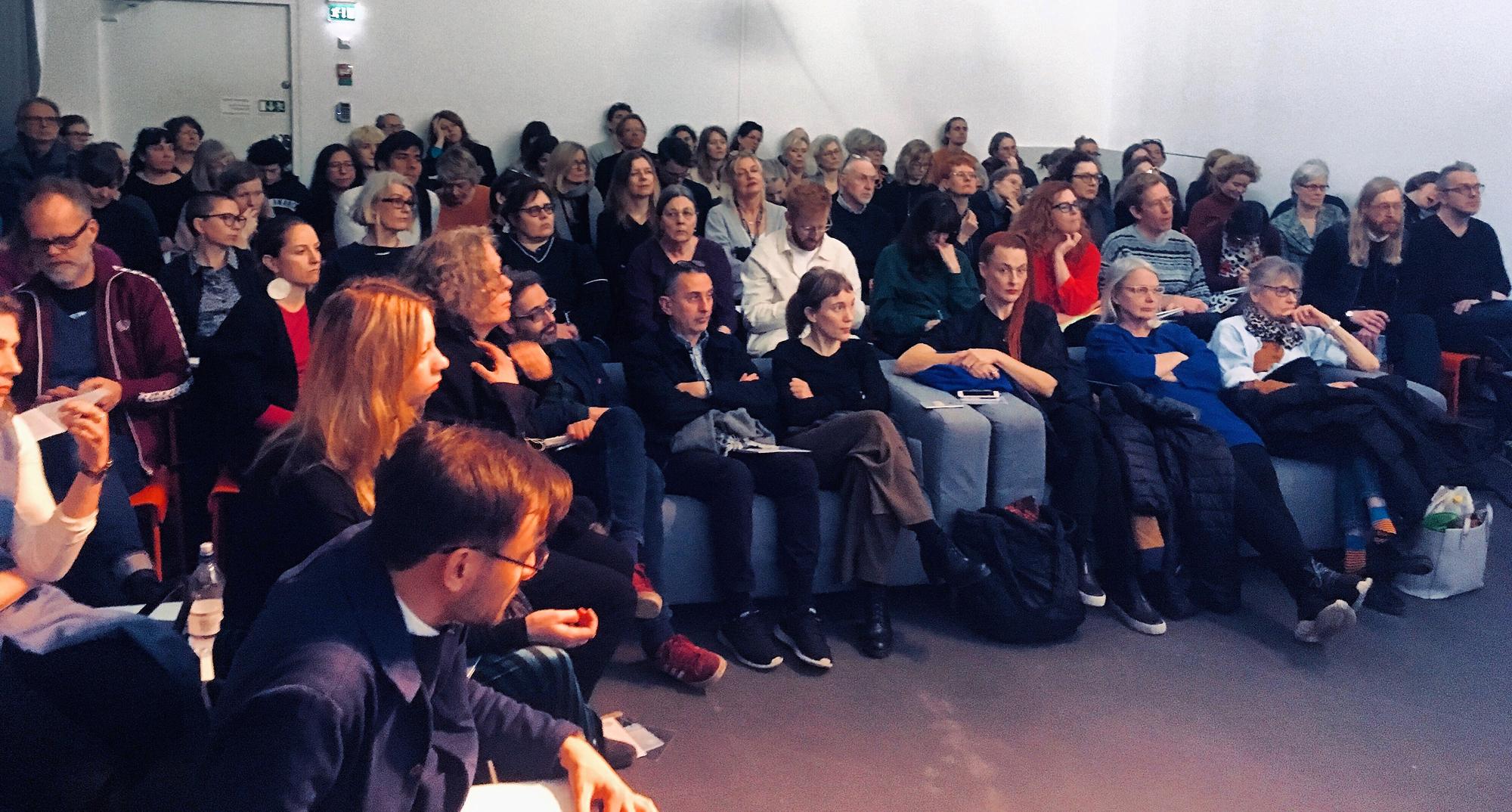 A large audience of adults seated in rows, facing forward in a modern room.