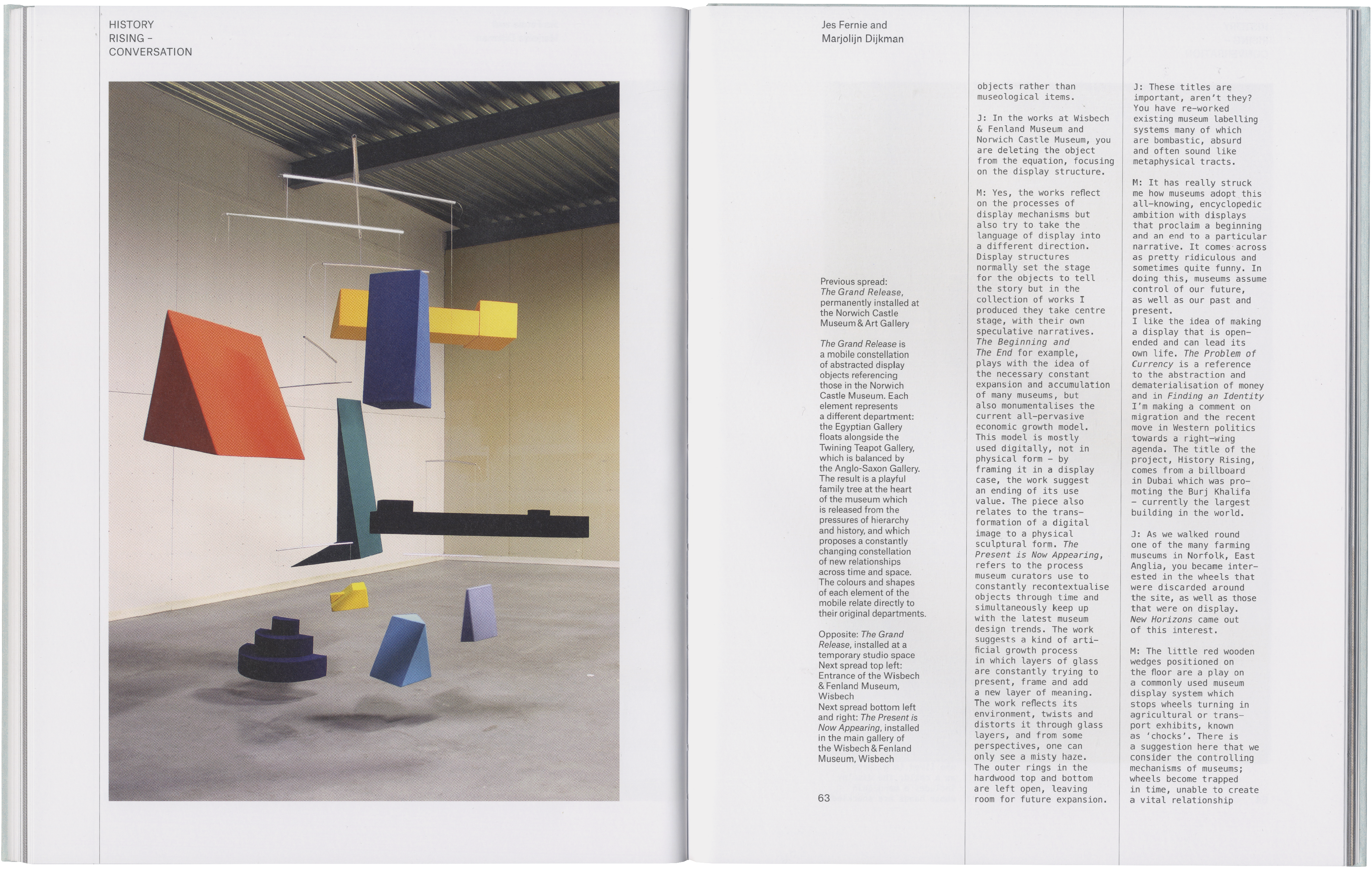 An abstract mobile sculpture made of red, blue, yellow, black, green, and purple geometric shapes suspended from the ceiling and arranged on the floor of a minimalist room.