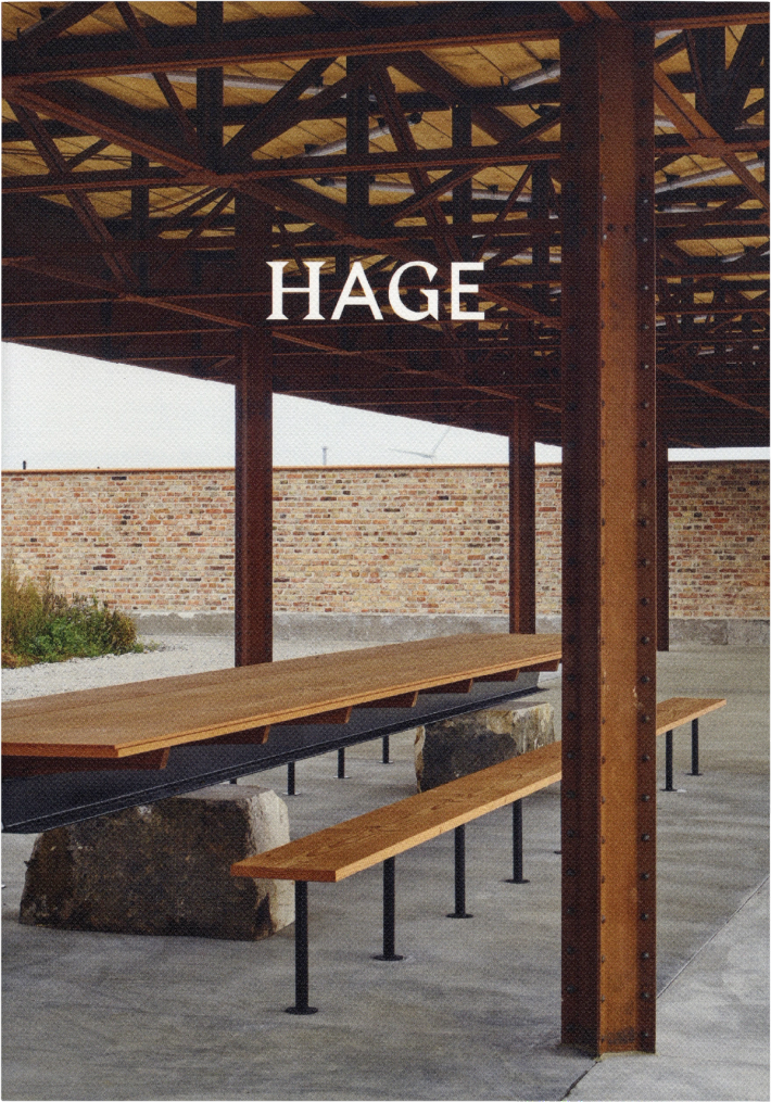 Outdoor pavilion with rusty metal roof, a long wooden table and bench supported by large stones, and a brick wall in the background. Text: HAGE.