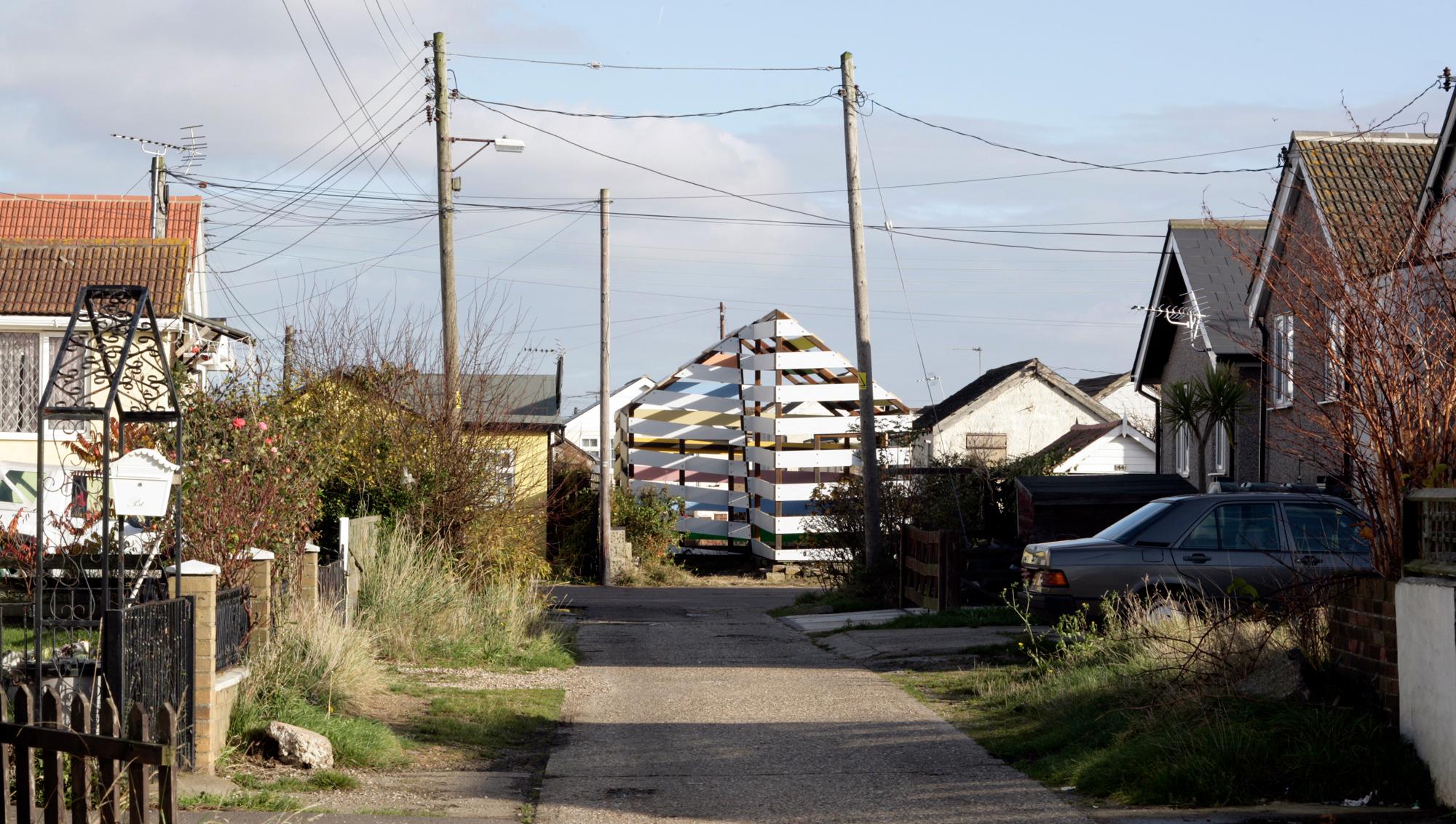 A narrow, overgrown road leads through a residential area towards a pyramid-shaped structure of stacked colorful boats under a clear sky with power lines.