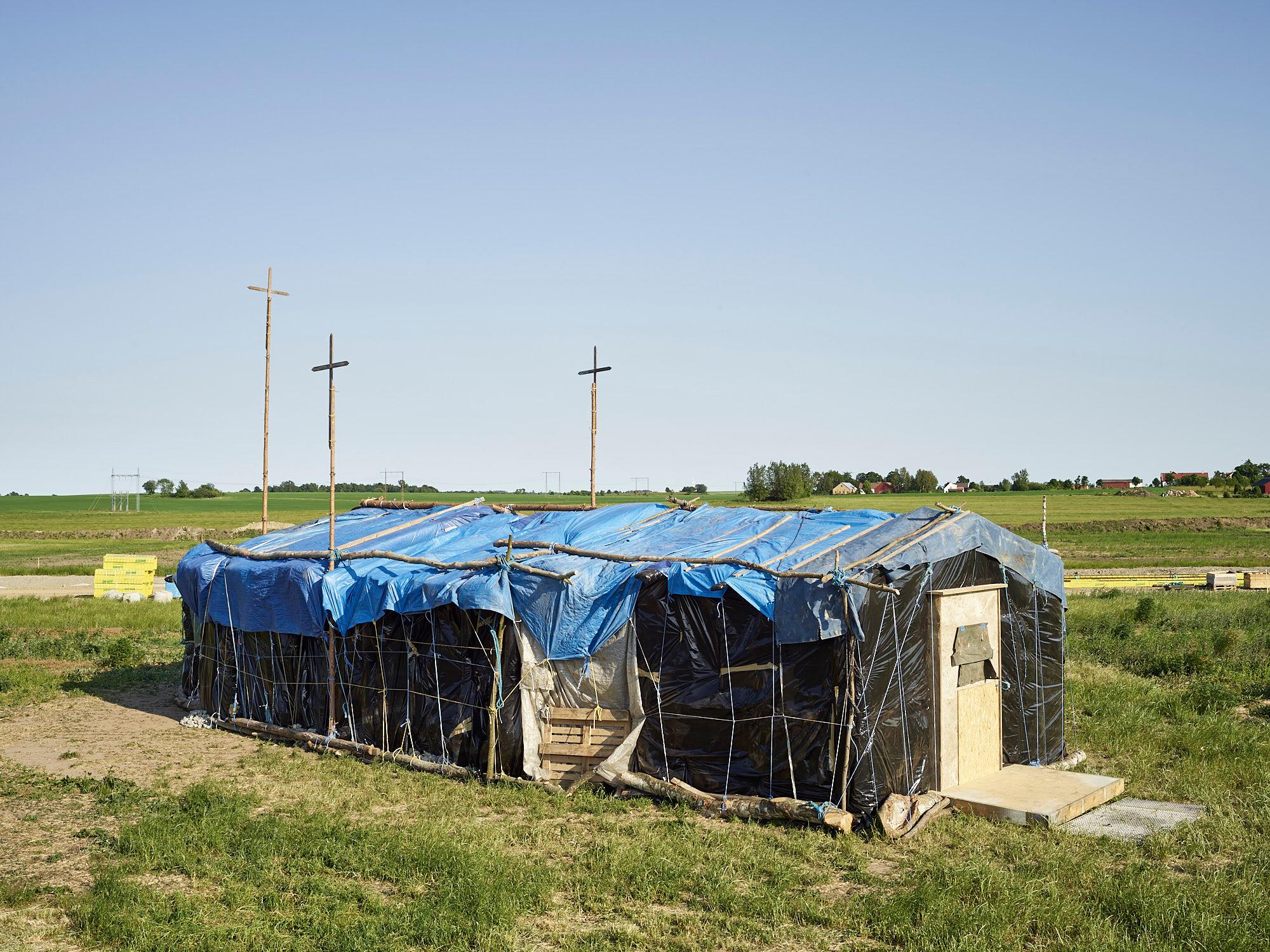 A makeshift building covered in black and blue tarps, topped with three wooden crosses, in a grassy field under a clear sky.