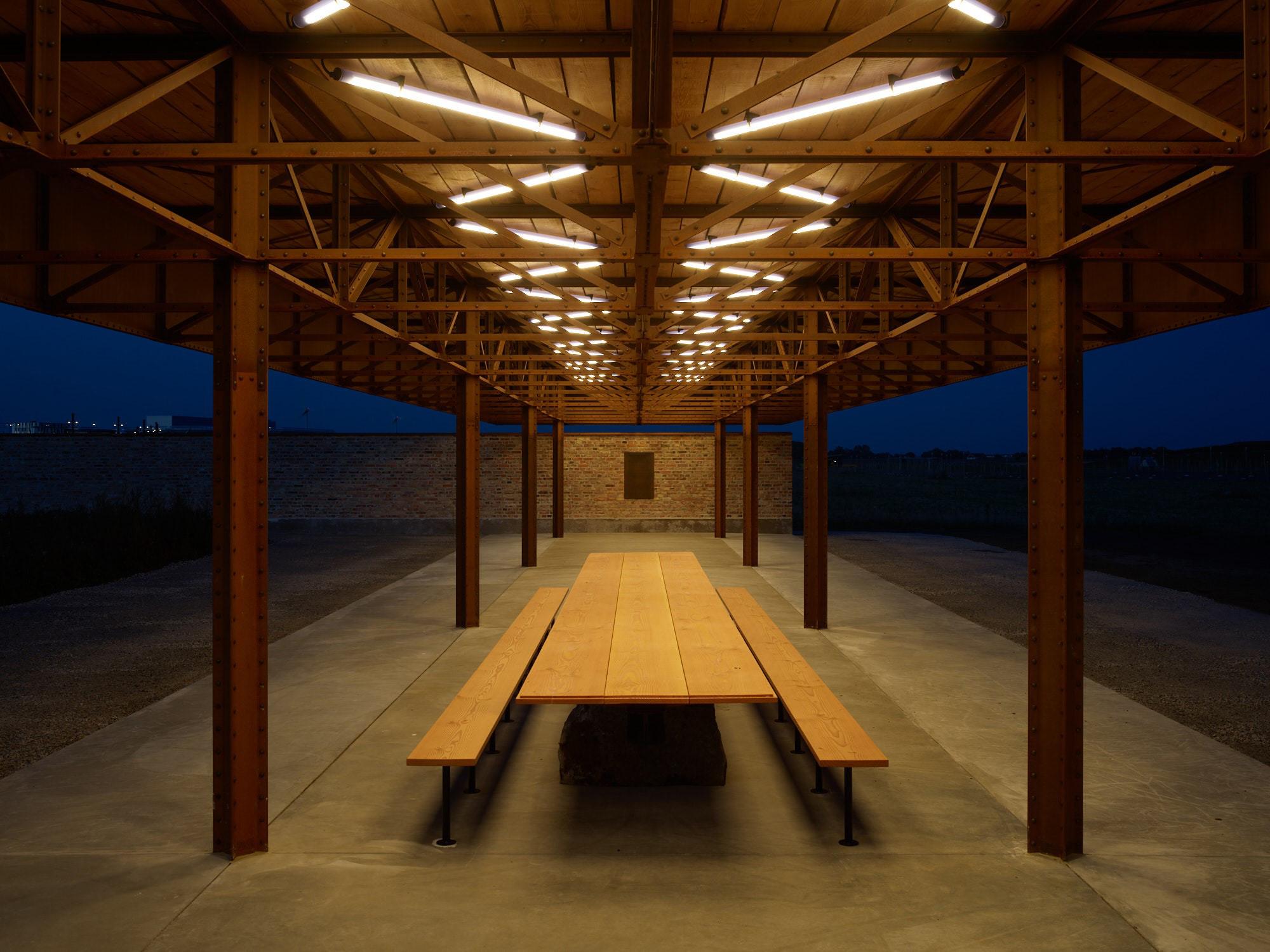 A long wooden pavilion with a picnic table and benches, brightly lit by overhead fluorescent lights at night.