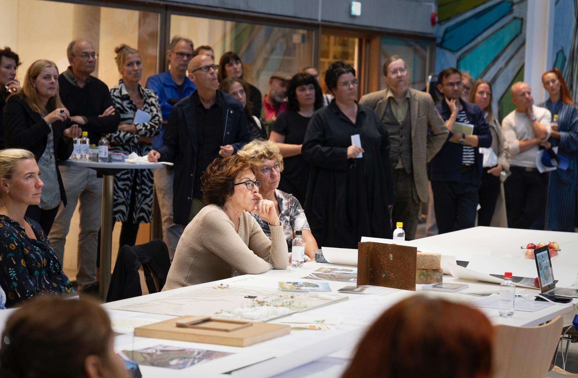 A group of people attending an indoor event, with some seated at a table covered in papers and models, and others standing and observing in the background.