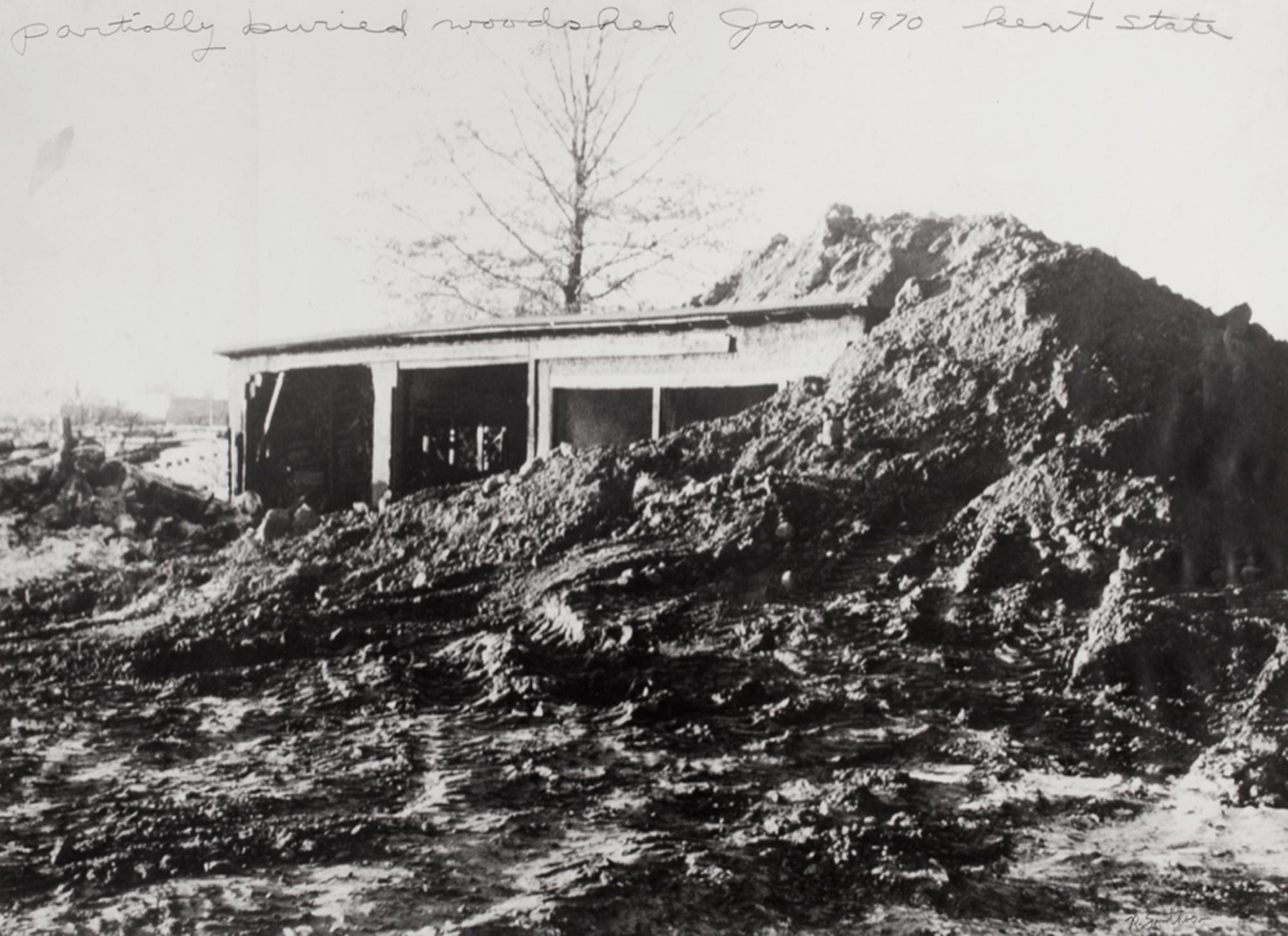 Black and white photo of a wooden shed partially buried by a large dirt mound on muddy ground at Kent State, January 1970.