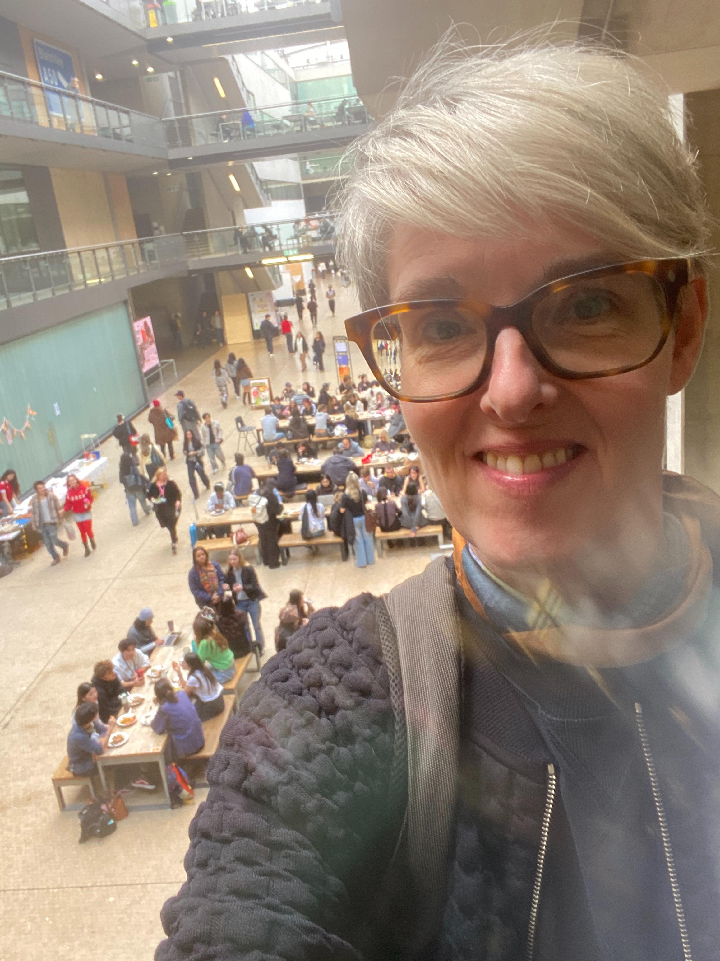 Selfie of a smiling woman with grey hair and glasses, looking down at a bustling indoor atrium filled with people.
