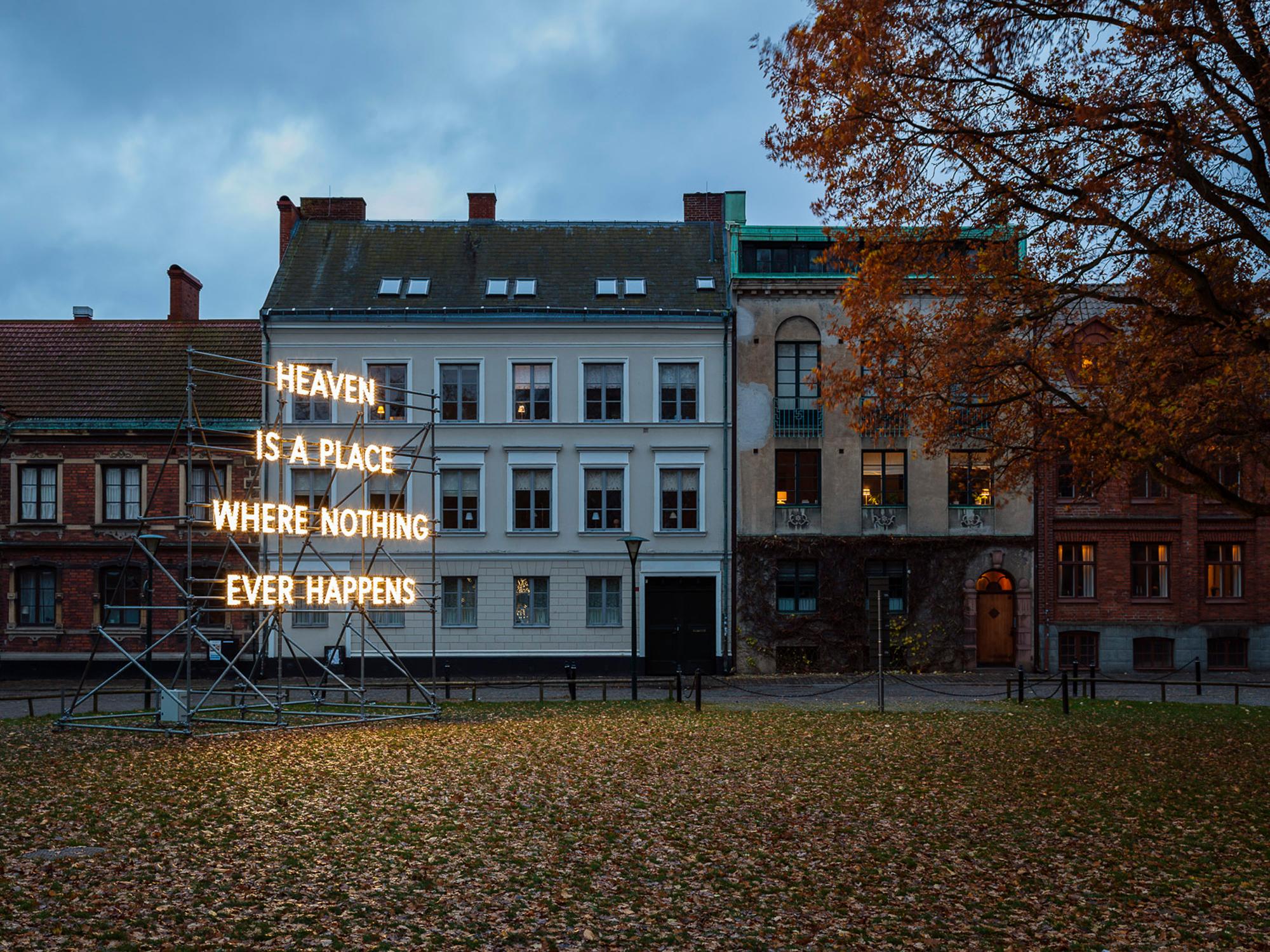 Large glowing neon sign reads "HEAVEN IS A PLACE WHERE NOTHING EVER HAPPENS" in a park with autumn leaves and historic buildings.