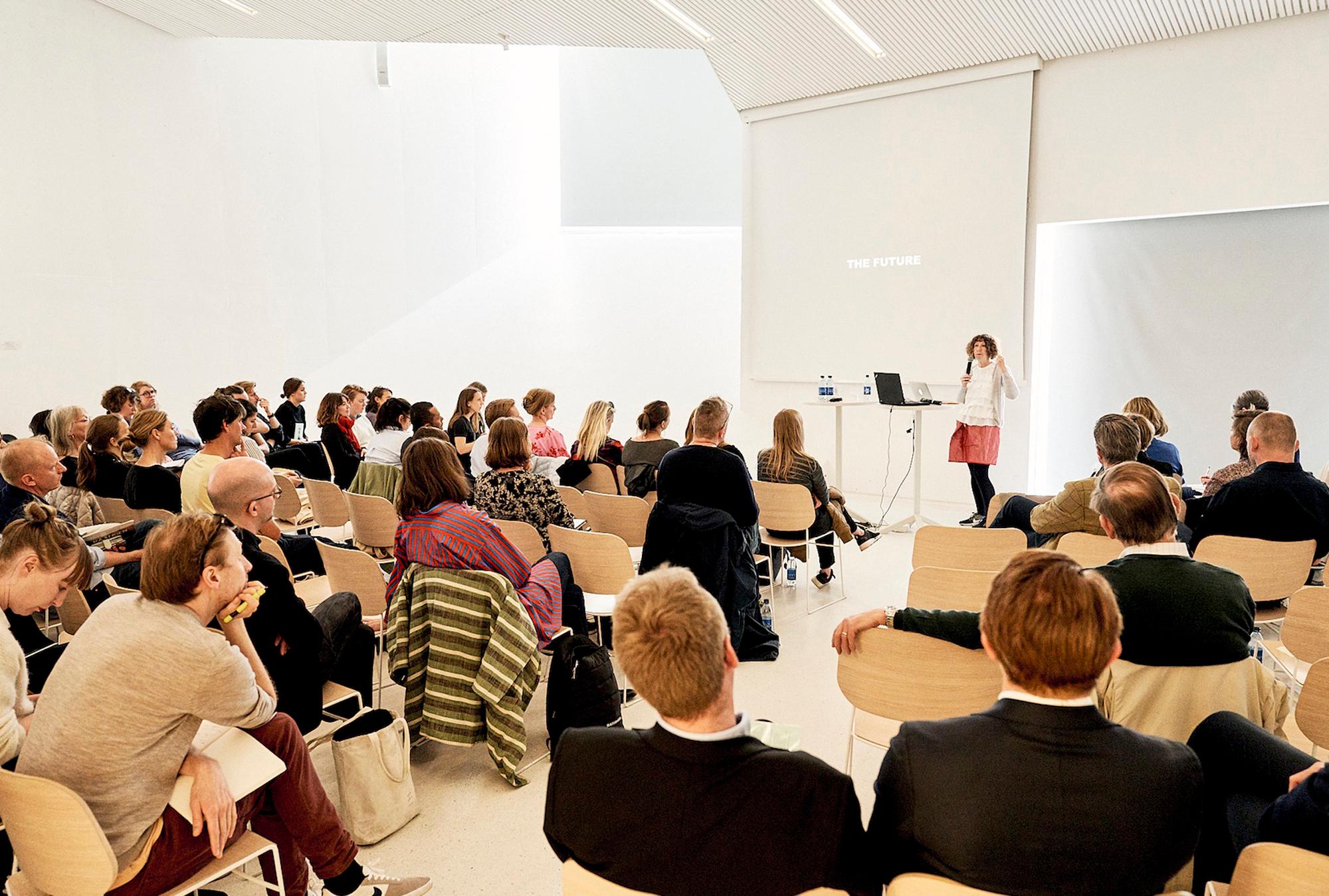 A woman presents "THE FUTURE" to a diverse audience in a modern lecture hall.