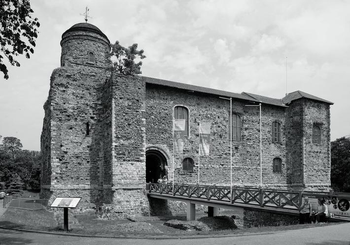 Black and white photo of a historic stone building with a prominent round tower, arched entrance, and a wooden bridge leading to it.