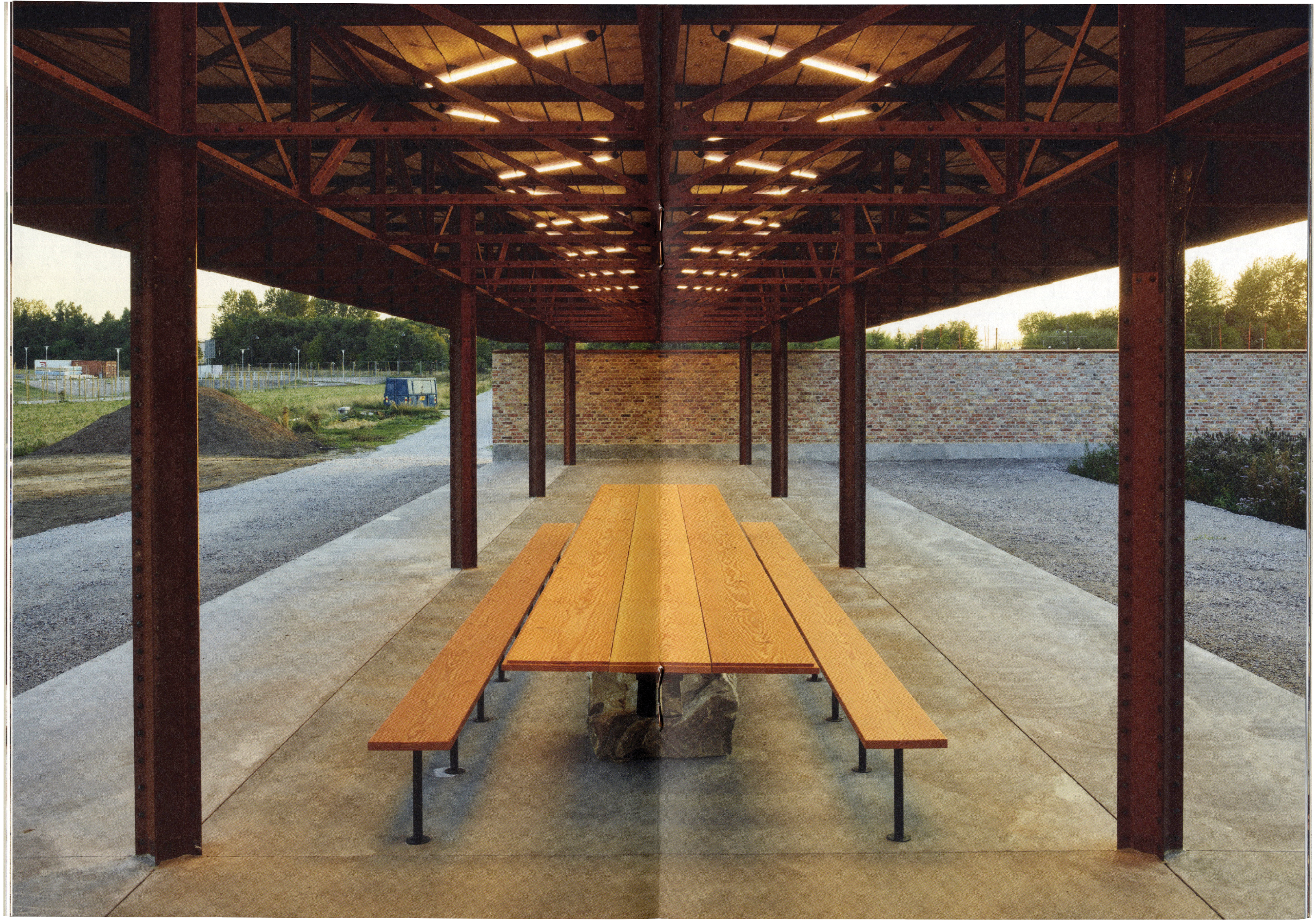 A long, open-air pavilion with a dark metal frame and illuminated ceiling, sheltering a wooden picnic table and benches. A gravel path, trees, and stone wall are in the background.