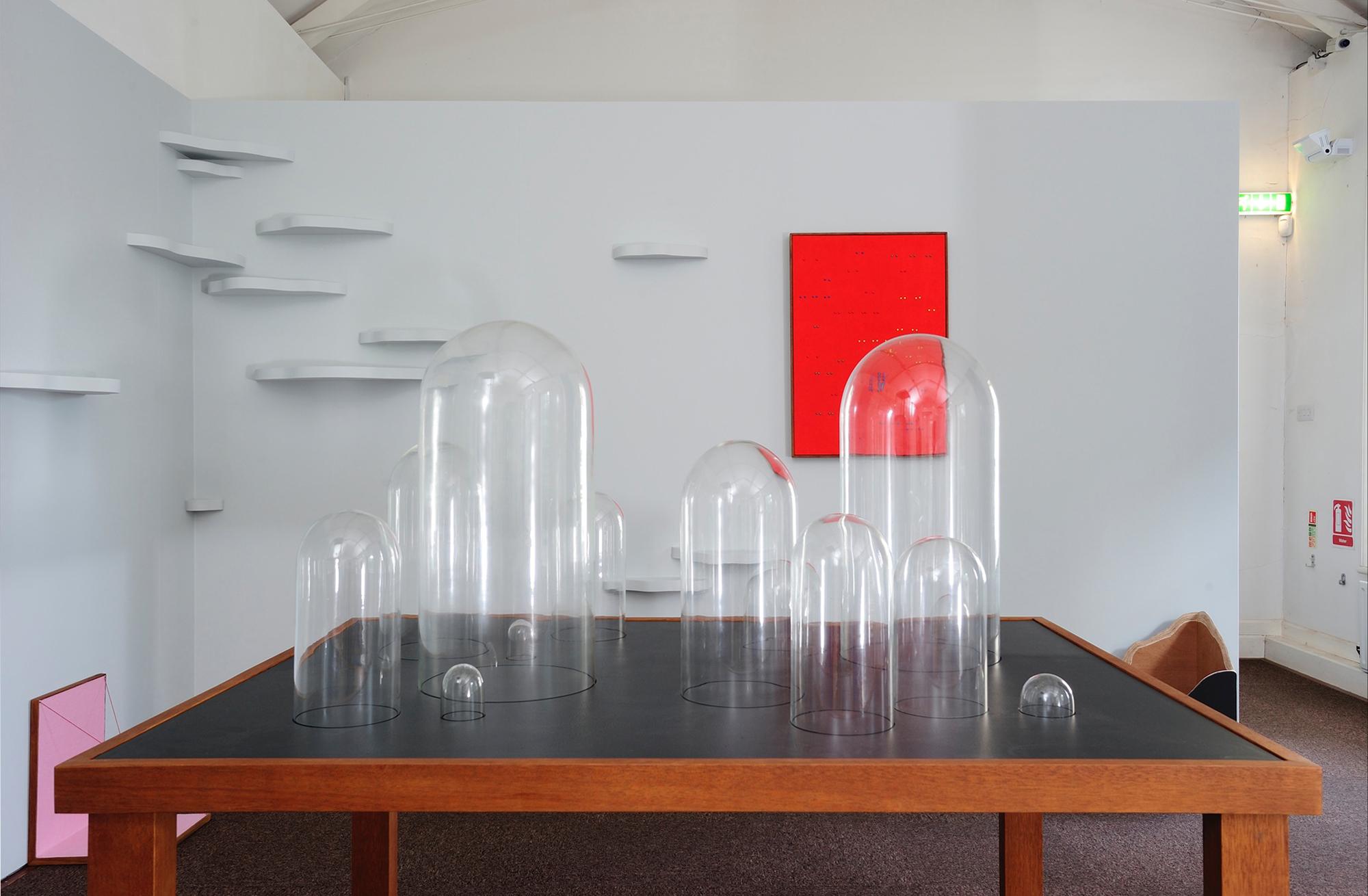 A contemporary art exhibit featuring clear glass cloches of various sizes on a table, backed by white minimalist shelves and a red rectangular artwork.