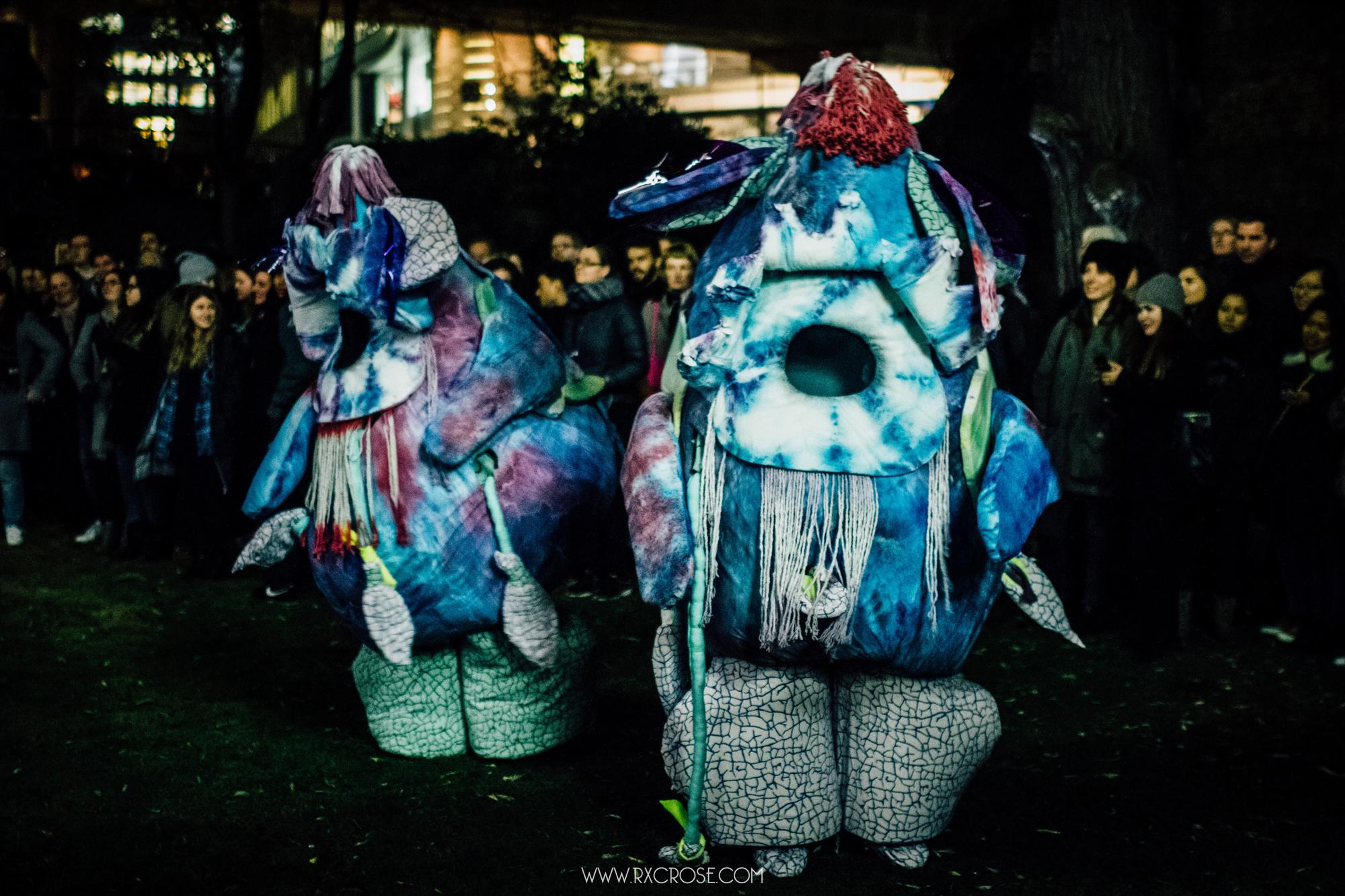 Two performers in large, abstract blue and purple tie-dye costumes, illuminated at night with a crowd watching.