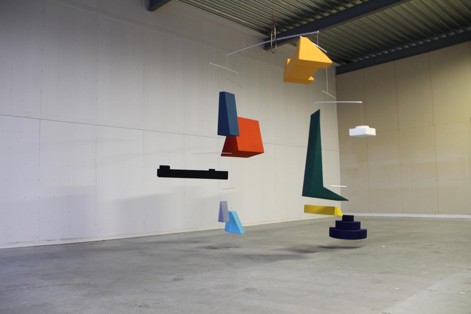A large mobile sculpture composed of various brightly colored geometric shapes hangs in a stark, empty gallery or industrial space.