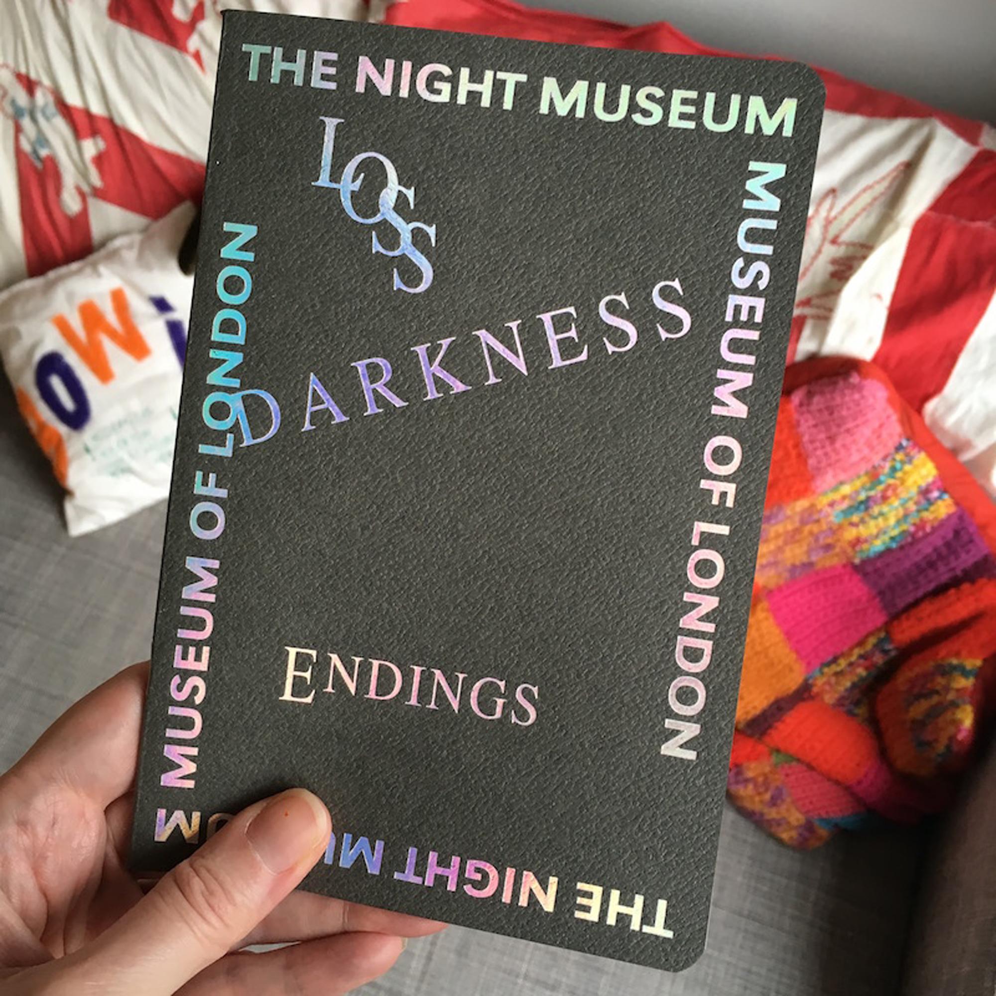 A hand holds a dark gray notebook with holographic text that reads "LOSS", "DARKNESS", "ENDINGS", "THE NIGHT MUSEUM", and "MUSEUM OF LONDON".