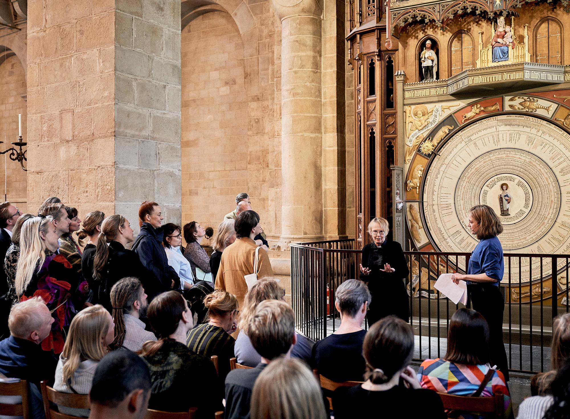 An audience listens to two women presenting in front of a grand astronomical clock inside a church.