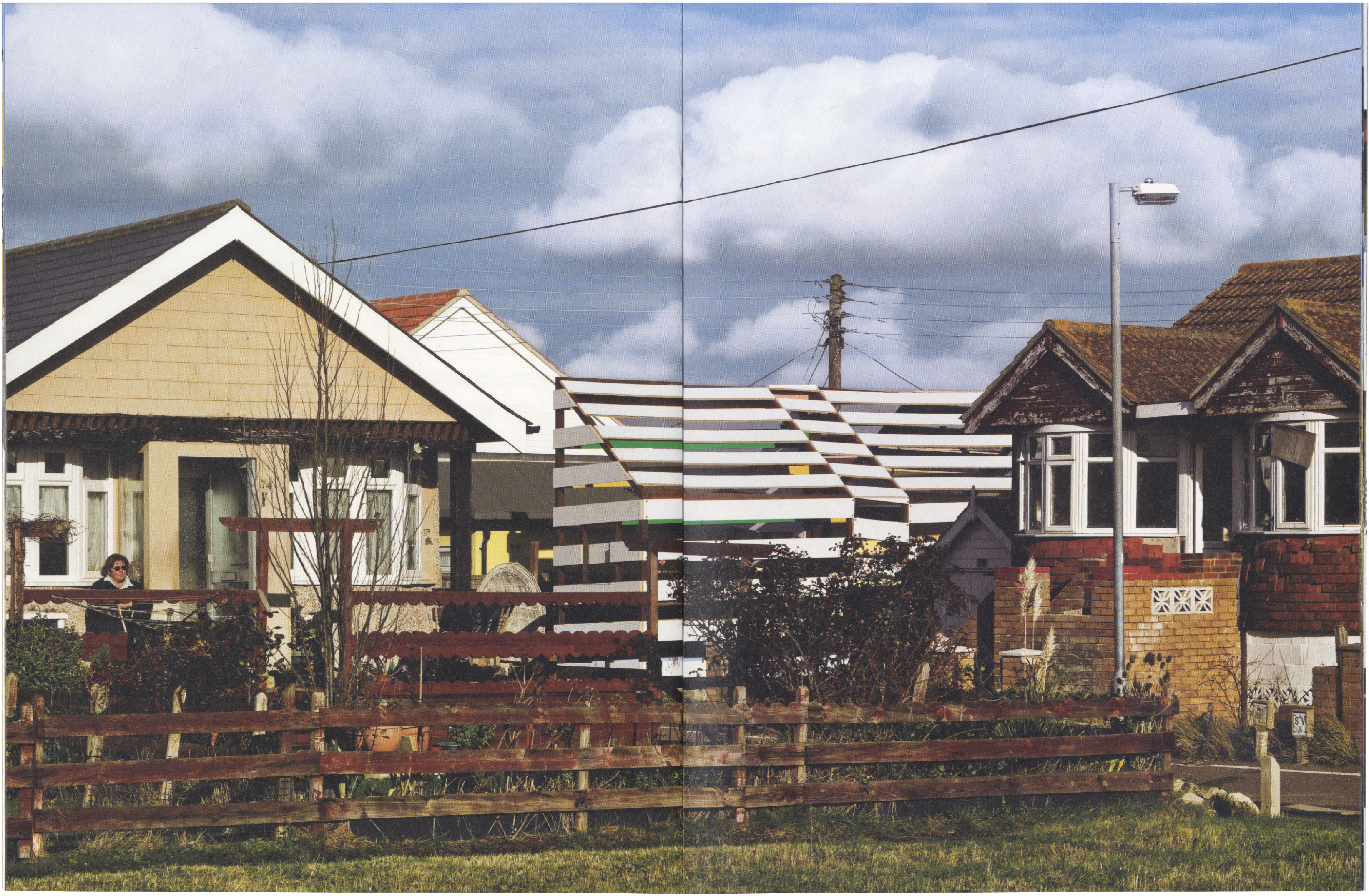 A suburban street scene with houses behind a wooden fence, featuring a prominent, horizontally striped structure that creates an optical illusion.